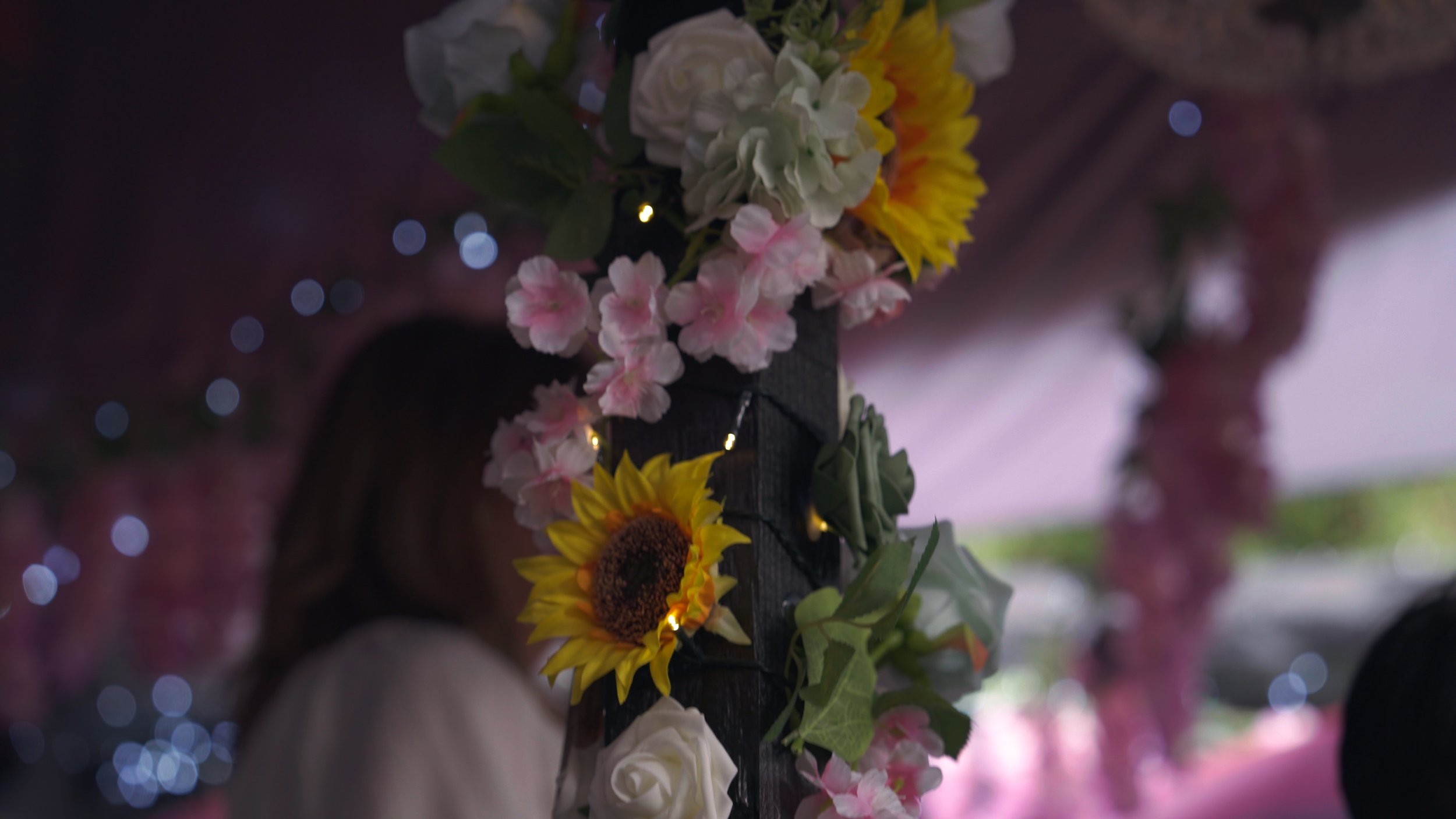 Close-up of a floral decoration with sunflowers, pink and white flowers, and green leaves, adorned with small string lights, with blurred background of a woman and a decorated venue.