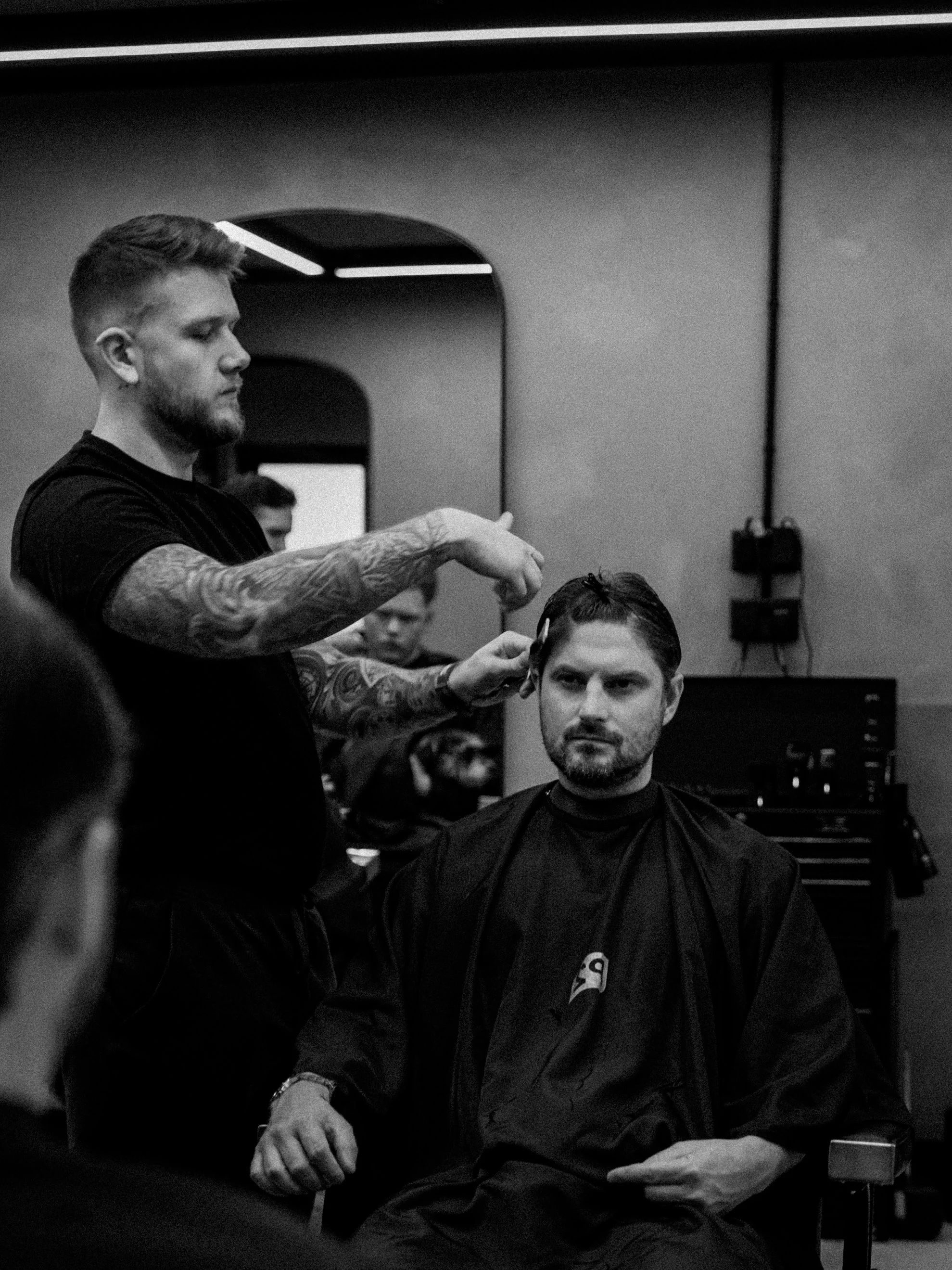 A man with a beard sitting in a barber's chair having his hair cut by a barber with tattoos on his arms, while other people are visible in the background.