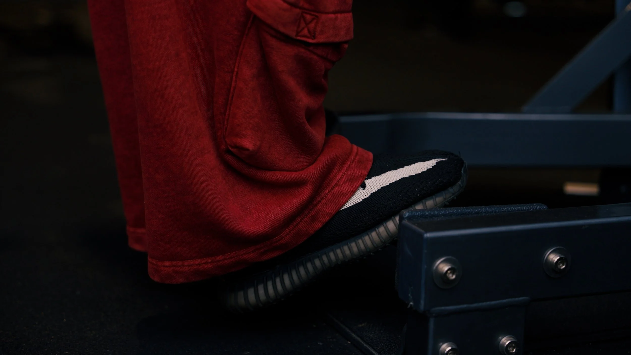 Close-up of a person's feet in black sneakers with white stripes, standing on a treadmill with a black surface and metal blue frame, wearing red sweatpants.