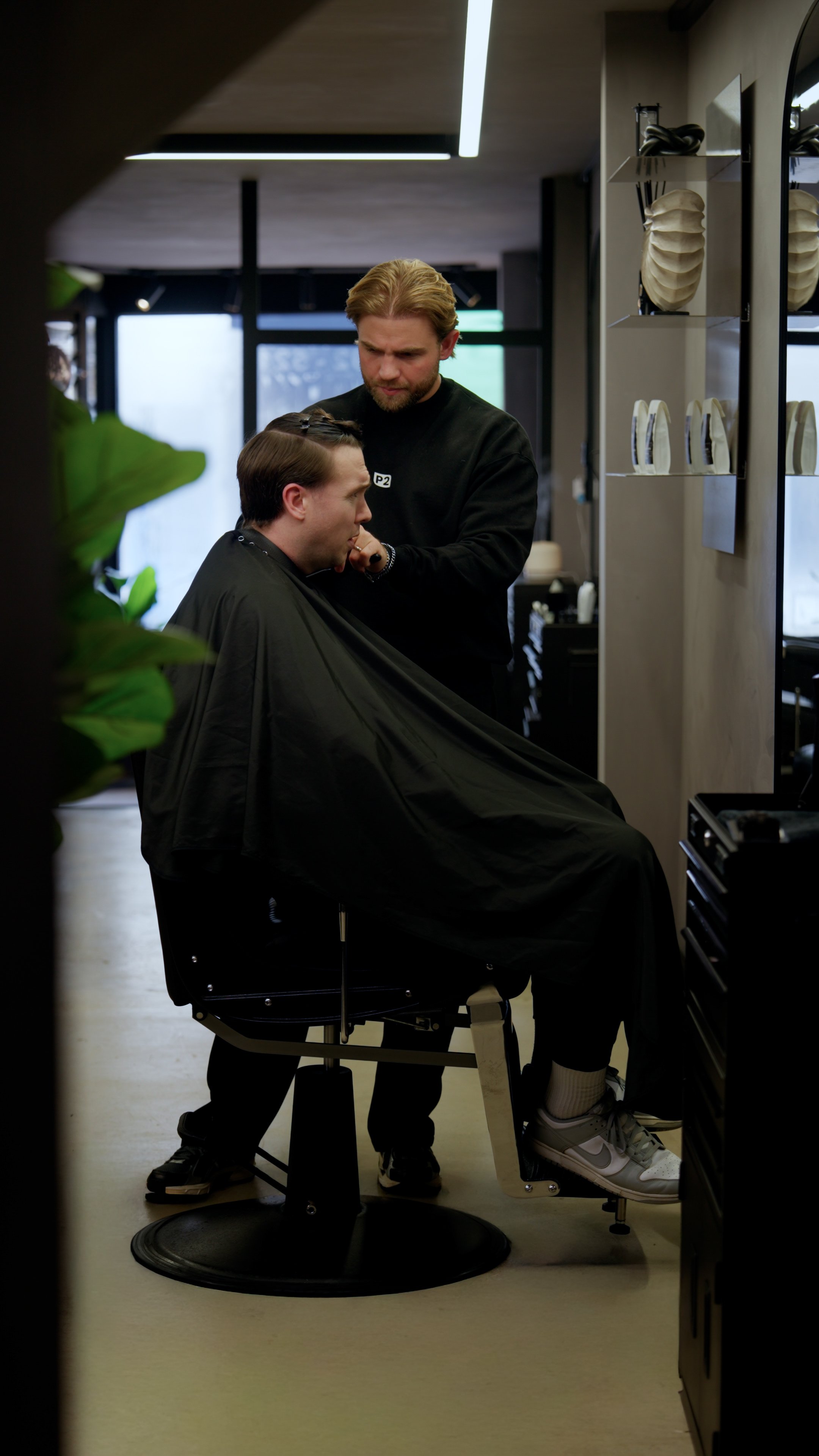 A man in a barber's chair getting a haircut from a barber in a modern salon, with salon tools and decor visible in the background.