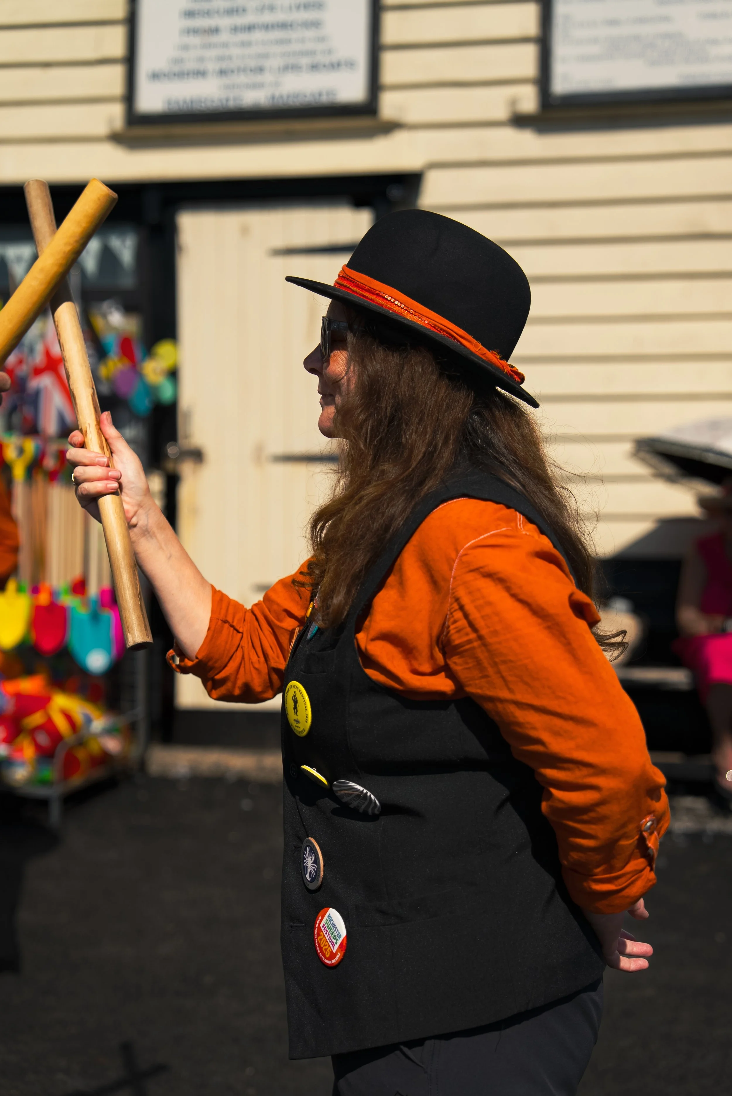 Woman wearing a black hat with red band, sunglasses, orange long-sleeve shirt, and a black vest with various buttons, holding two wooden sticks, standing outdoors near a building with colorful items on display.