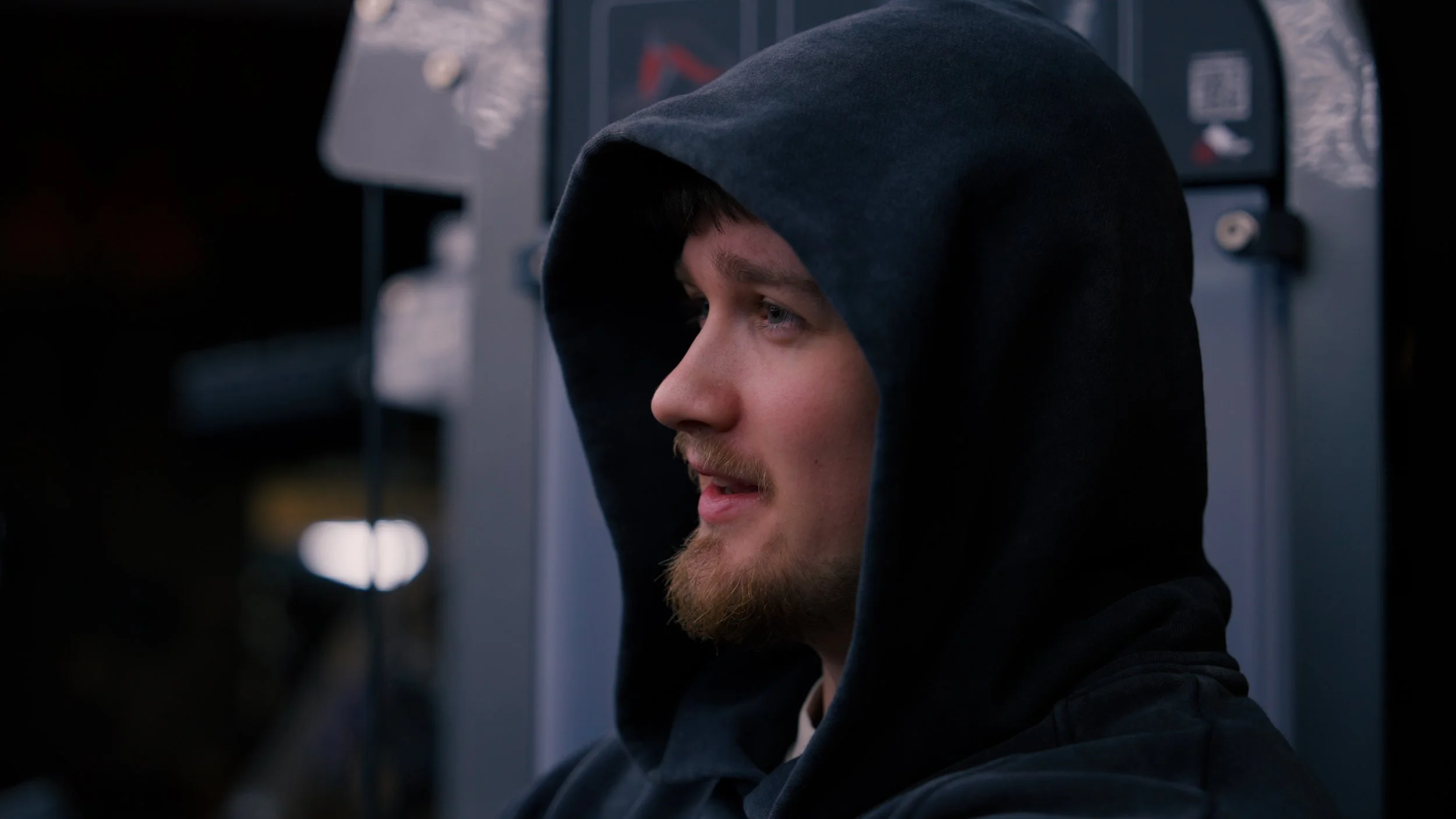A young man with a beard and blue eyes wearing a black hoodie with the hood over his head.