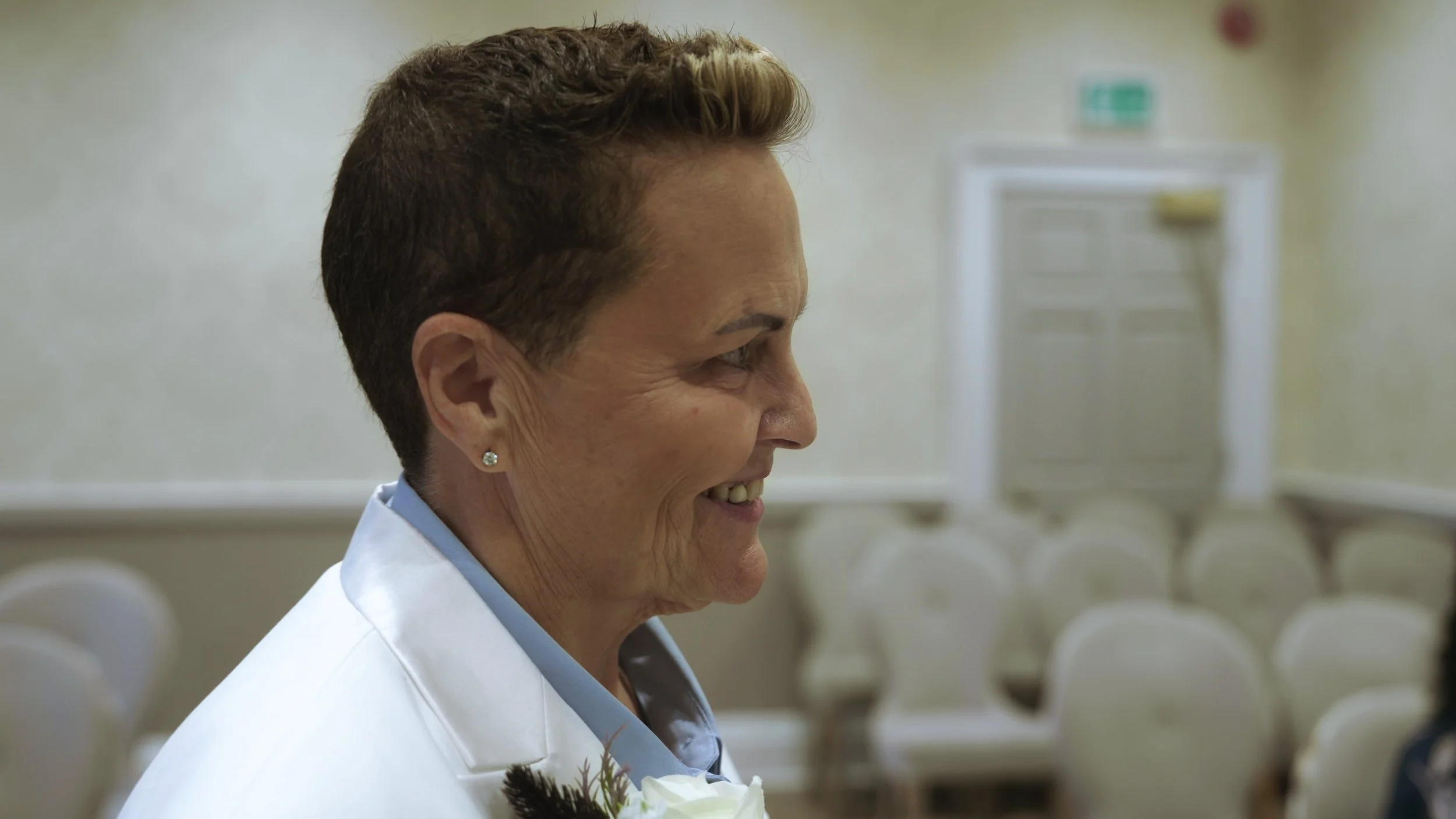 Profile of a smiling woman with short, styled hair, wearing a white coat and a small diamond earring, indoors with beige walls and rows of empty chairs in the background.