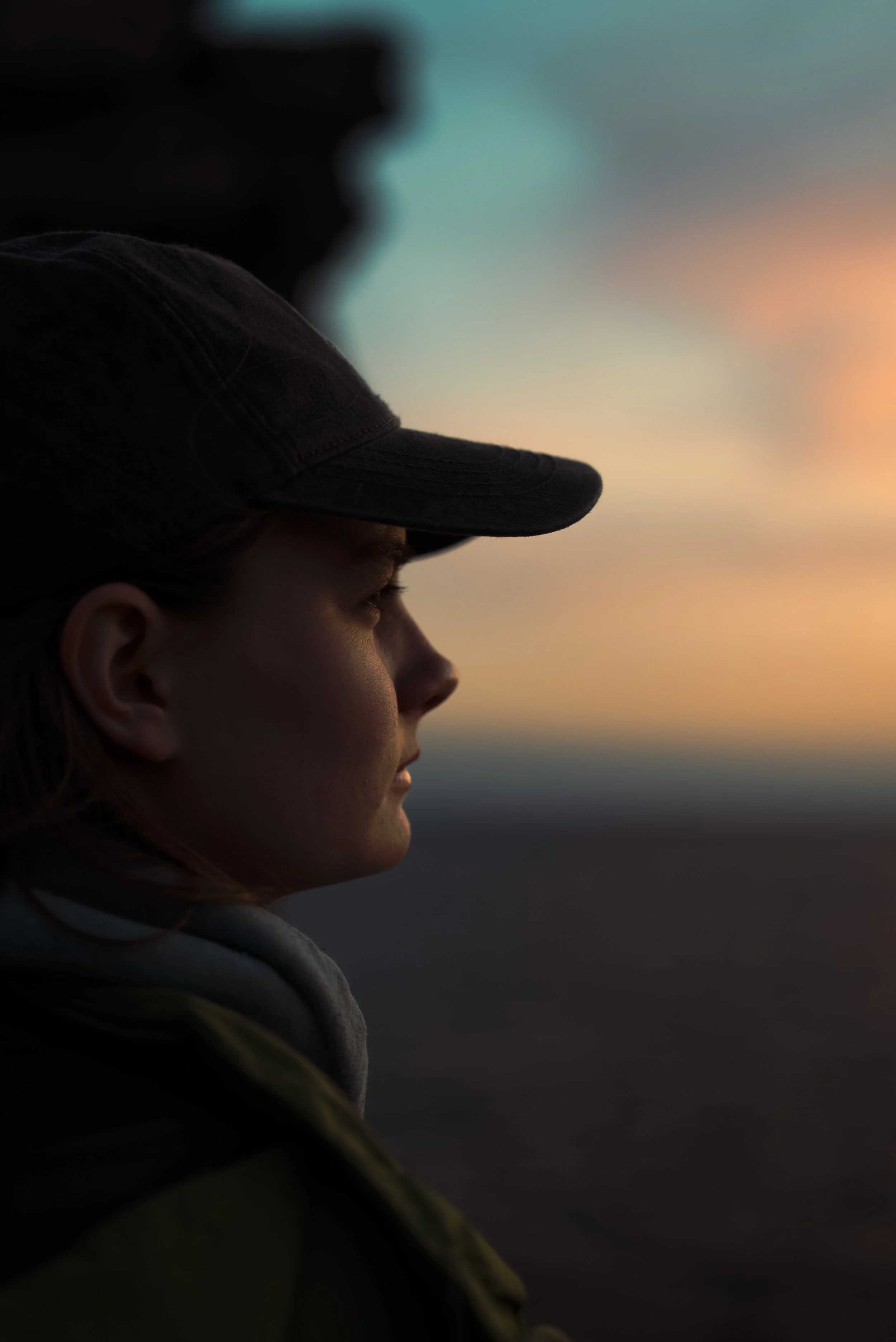Side profile of a woman wearing a baseball cap, looking at a sunset or sunrise sky with clouds.