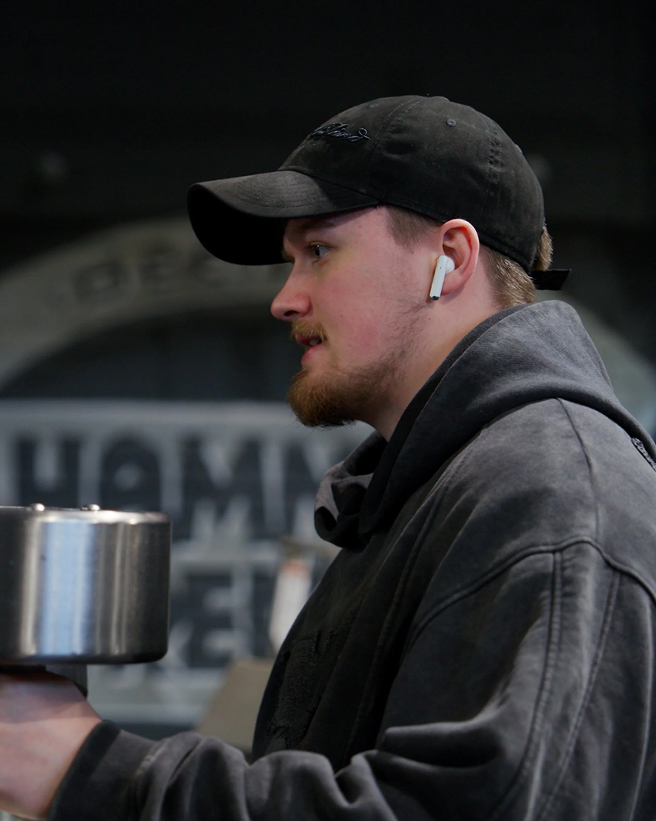 A man with a beard and mustache wearing a baseball cap, wireless earbuds, and a dark hoodie, holding a weight in a gym.