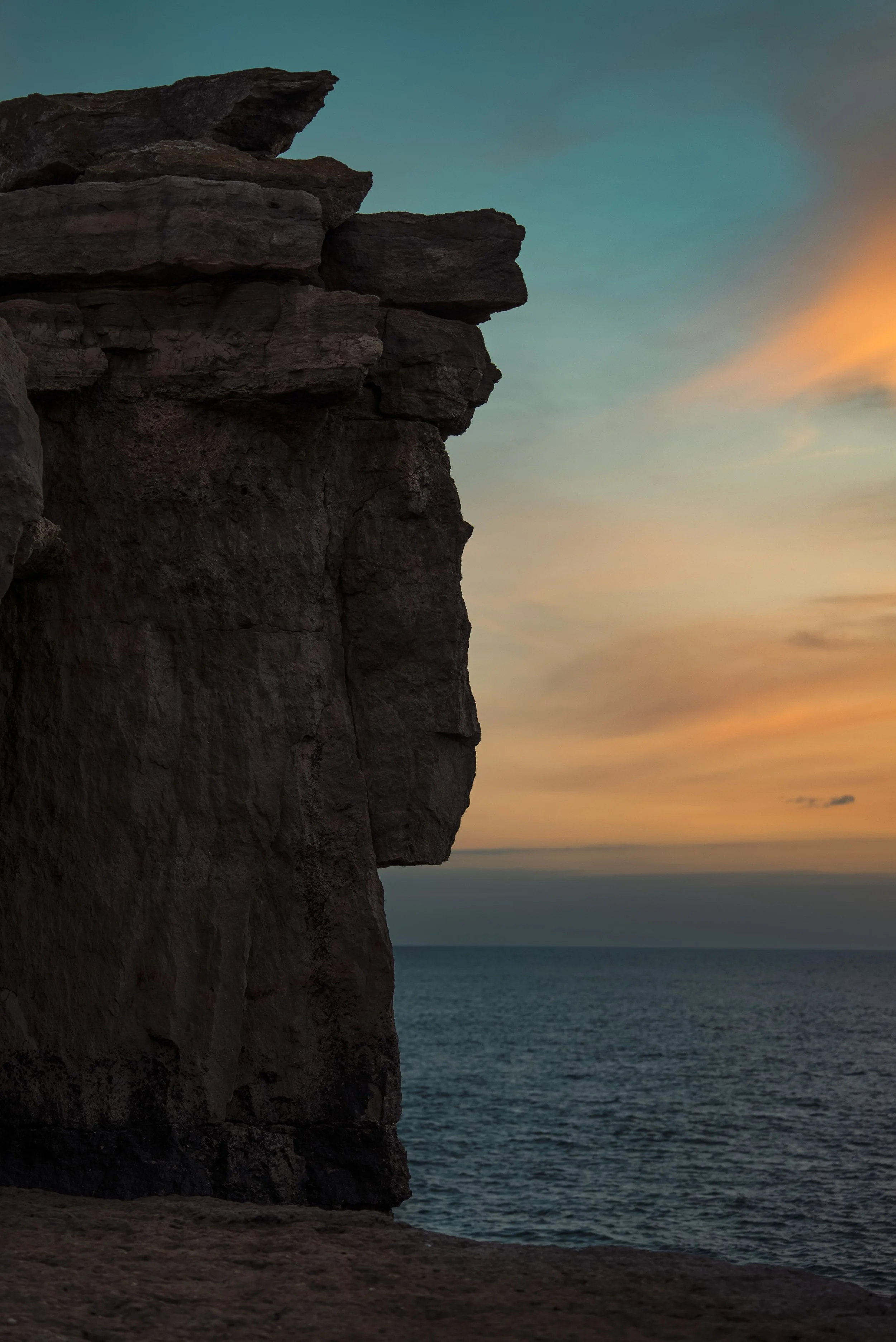 A rugged cliff face near the ocean during sunset, with a colorful sky of soft pastel hues and calm water.