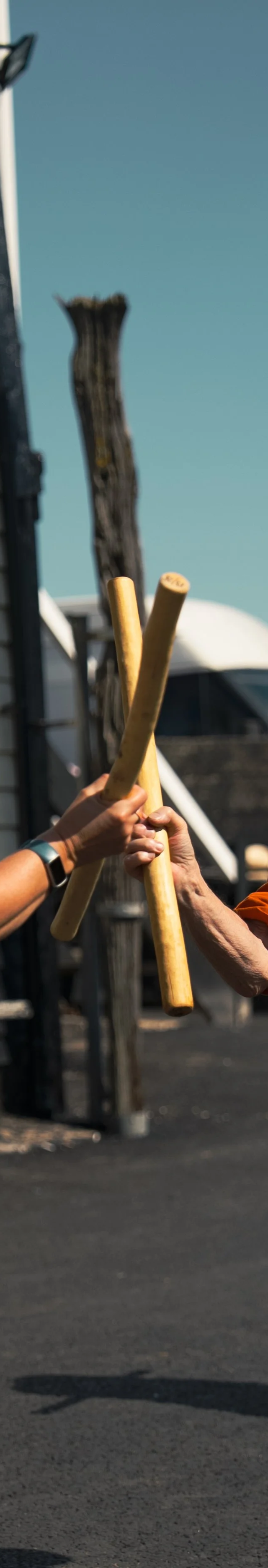 Two people holding sticks or batons, exchanging a high five outdoors on a paved surface with a wooden pole and a vehicle in the background.