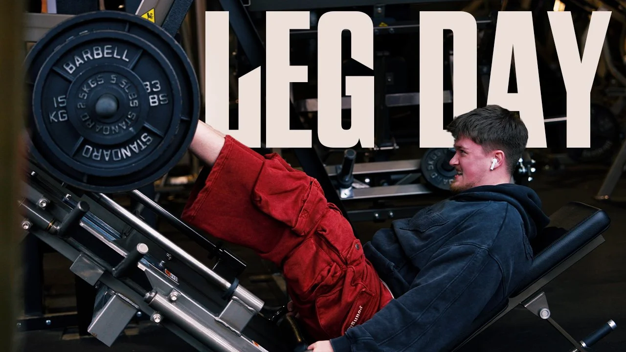 A man lying on a workout bench lifting a weight with one leg, wearing red pants and a dark hoodie in a gym, with the word 'LEG DAY' overlaid.