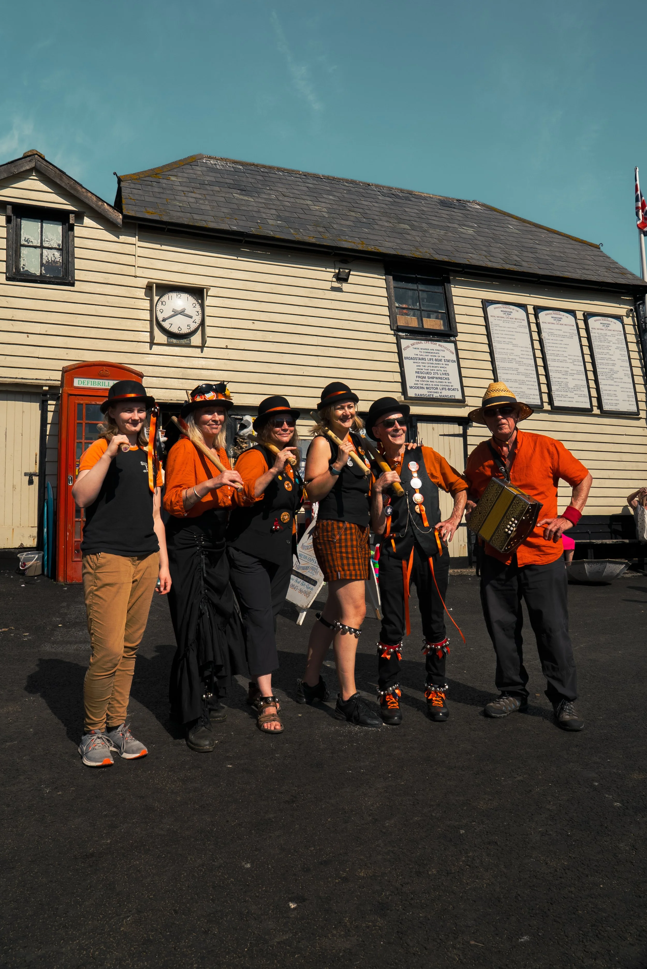 Six people dressed in black and orange costumes posing in front of a wooden building, some holding musical instruments, with a clock and informational signs behind them.