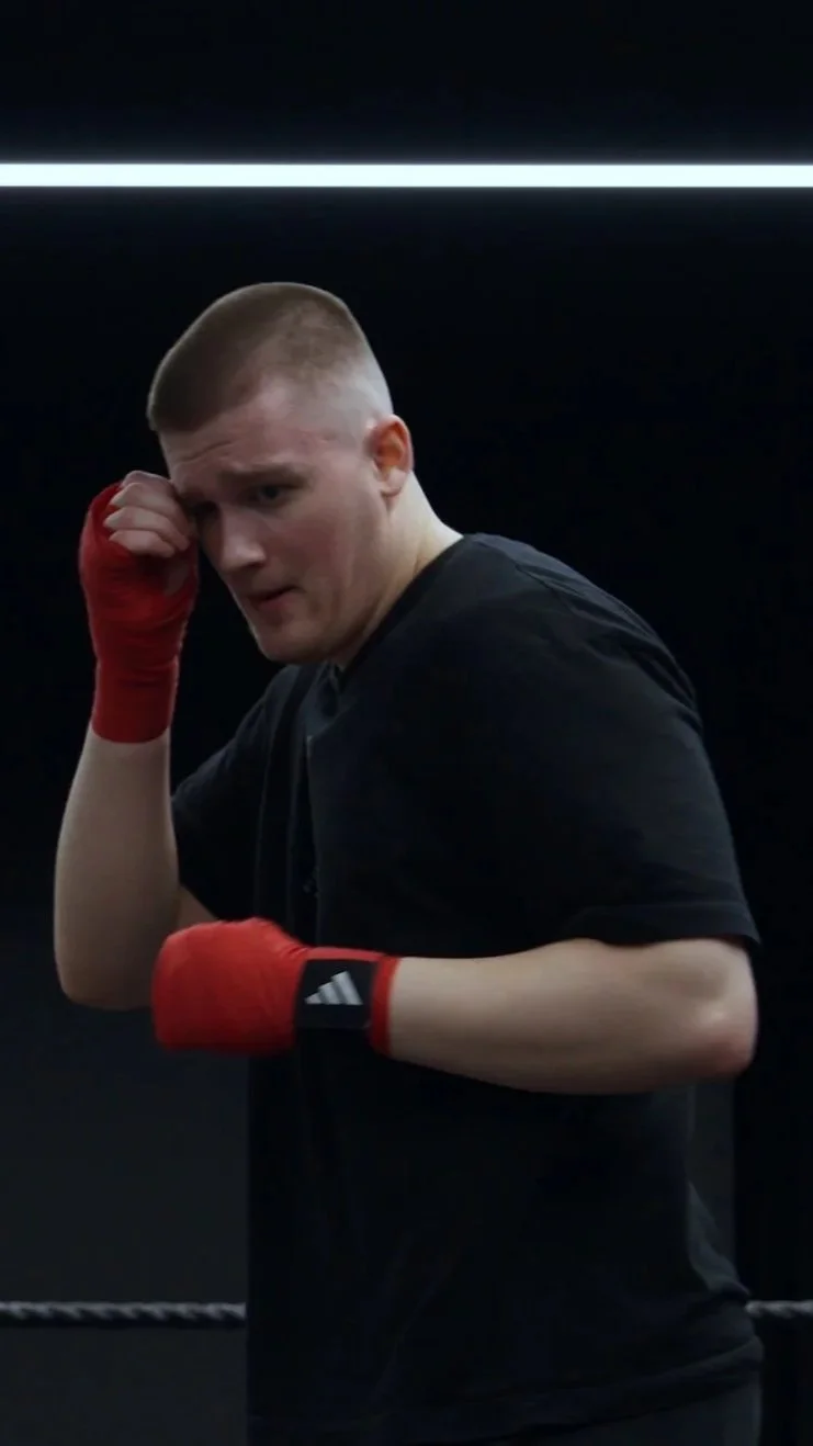 A young man with short blond hair wearing a black shirt and red boxing gloves, standing in a boxing gym with black walls and a bright strip of light above.