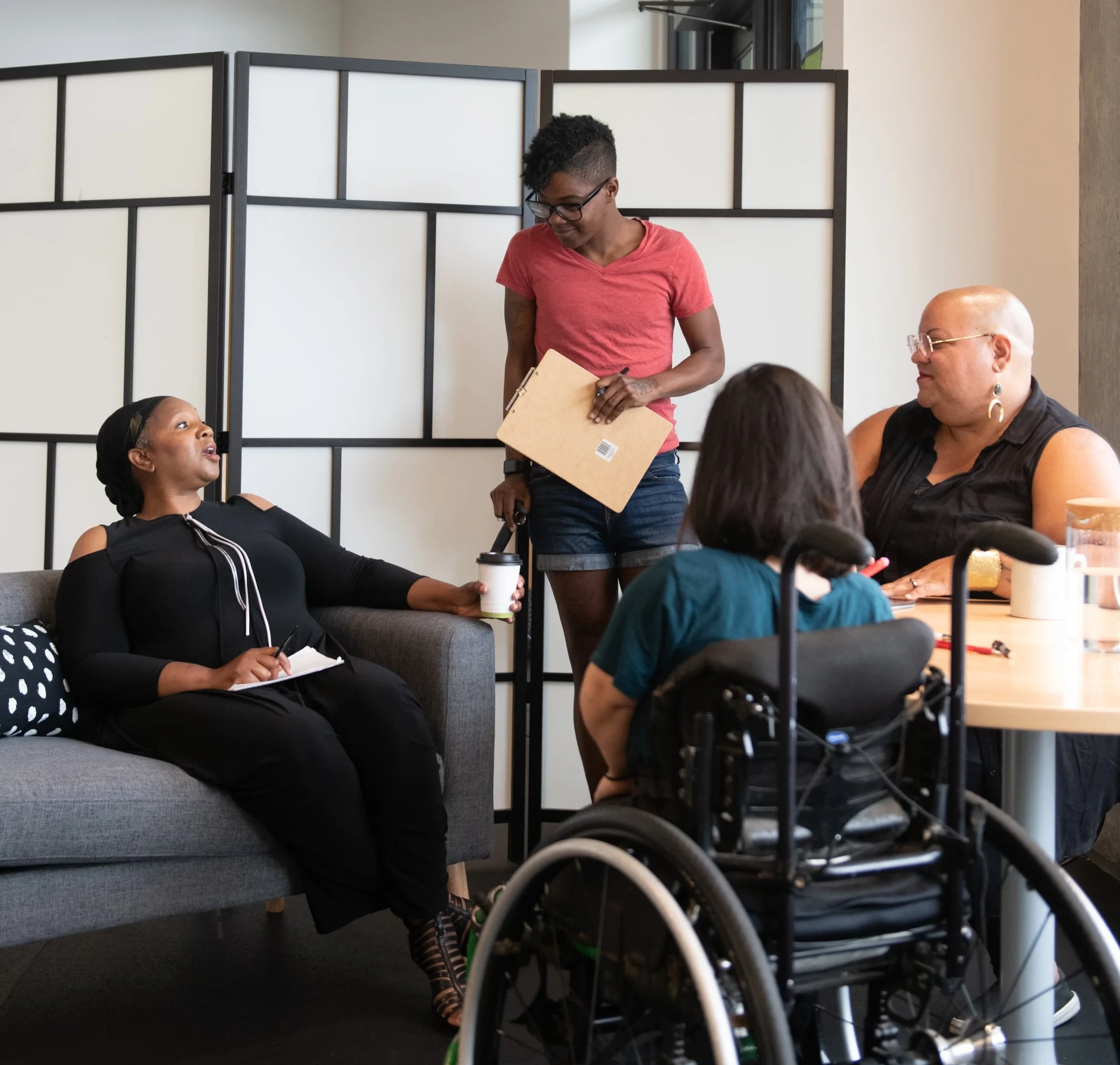 Group of four diverse women in a discussion in a modern office, one woman in a wheelchair, another woman standing with a clipboard, and two women sitting on a sofa and at a table.