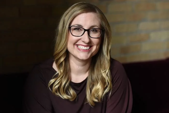 Smiling woman wearing glasses and a maroon shirt standing in front of a brick background.