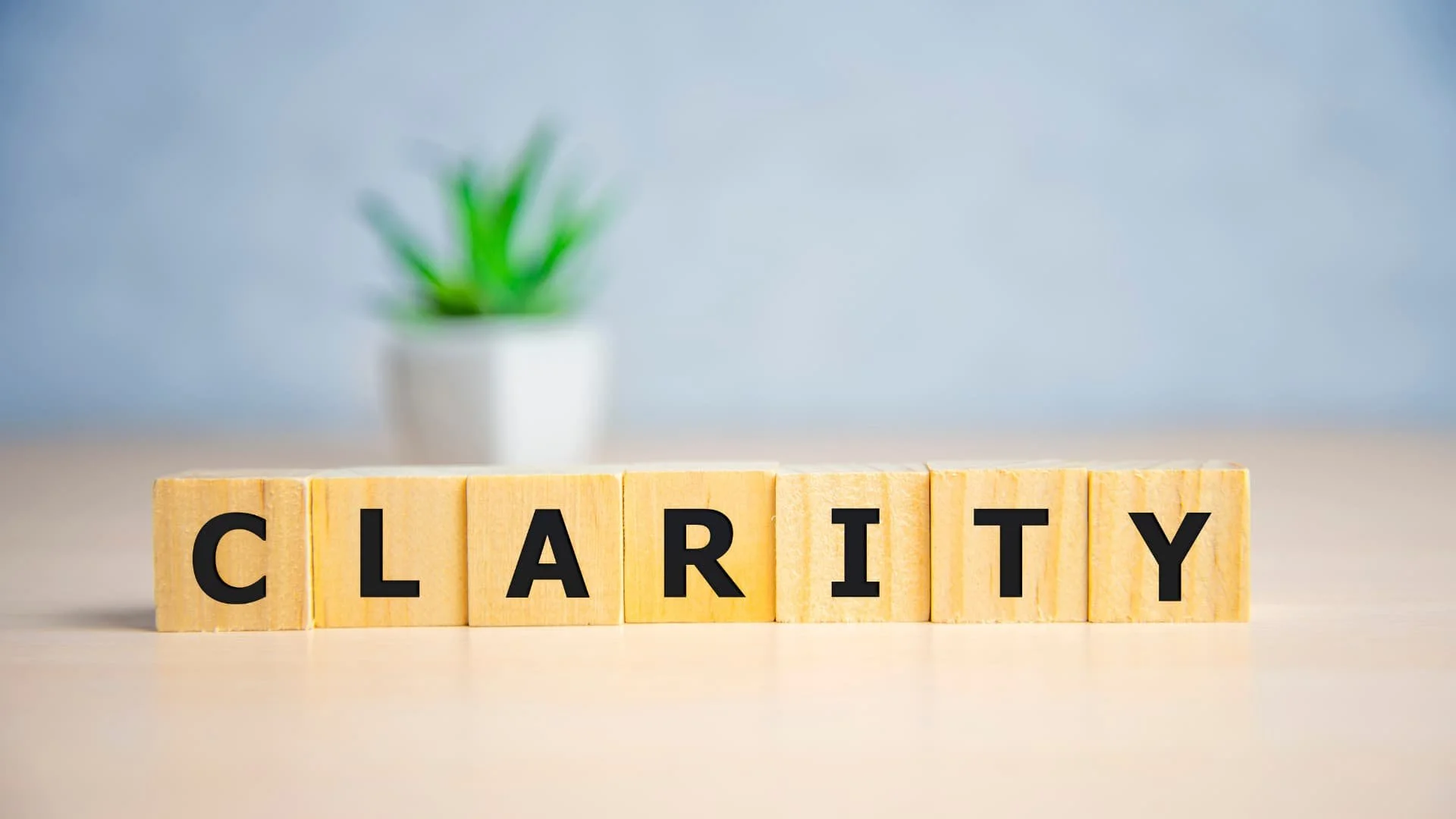 Wooden blocks spelling out 'CLARITY' with a blurred green plant in a white pot in the background.