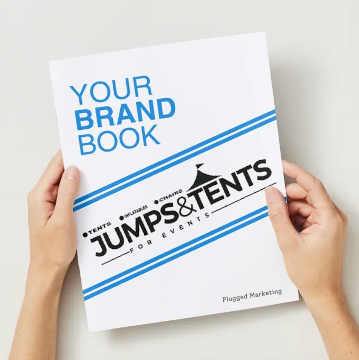 Person holding a white booklet titled 'Your Brand Book' with blue and black text about jumpers and tents for events, manufactured by Plugged Marketing.