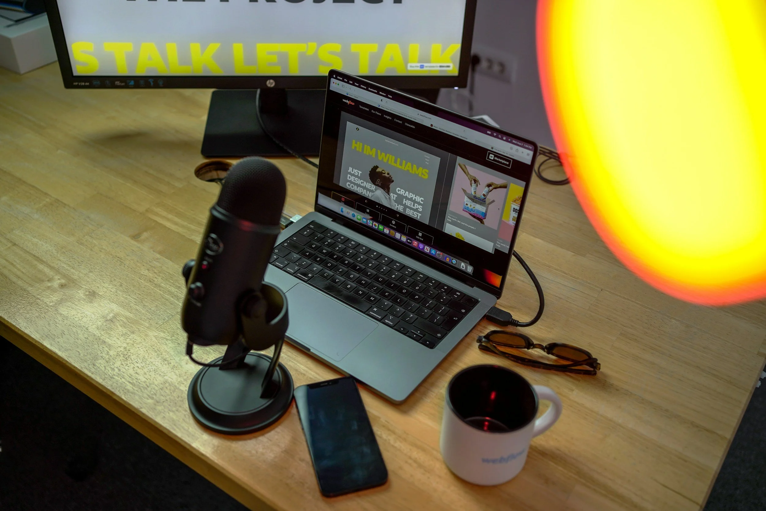 Work desk with a microphone, a smartphone, a white mug with black coffee, sunglasses, and a laptop displaying a webpage, with a monitor in the background showing the phrase 'STALK LET'S TALK' in bright yellow text.