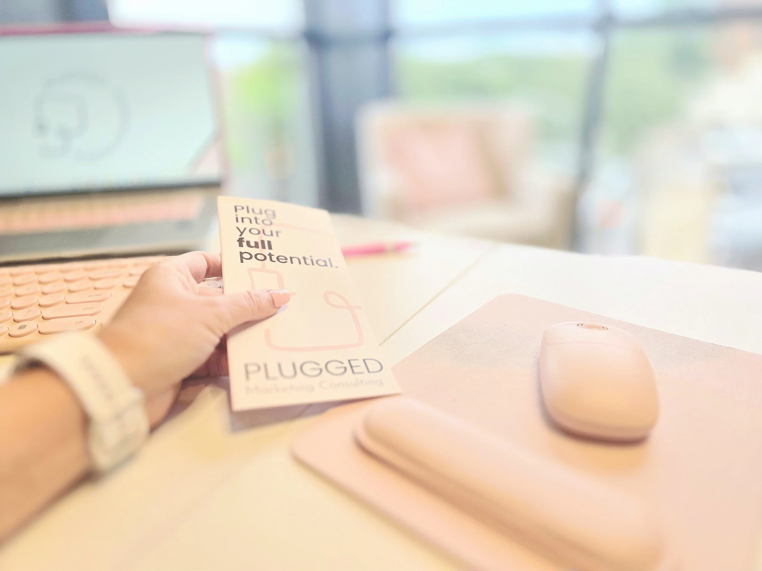 A person holding a pamphlet that says 'Plug into your full potential' at a workstation with a pink computer mouse on a pink mouse pad, a monitor, and a window in the background.