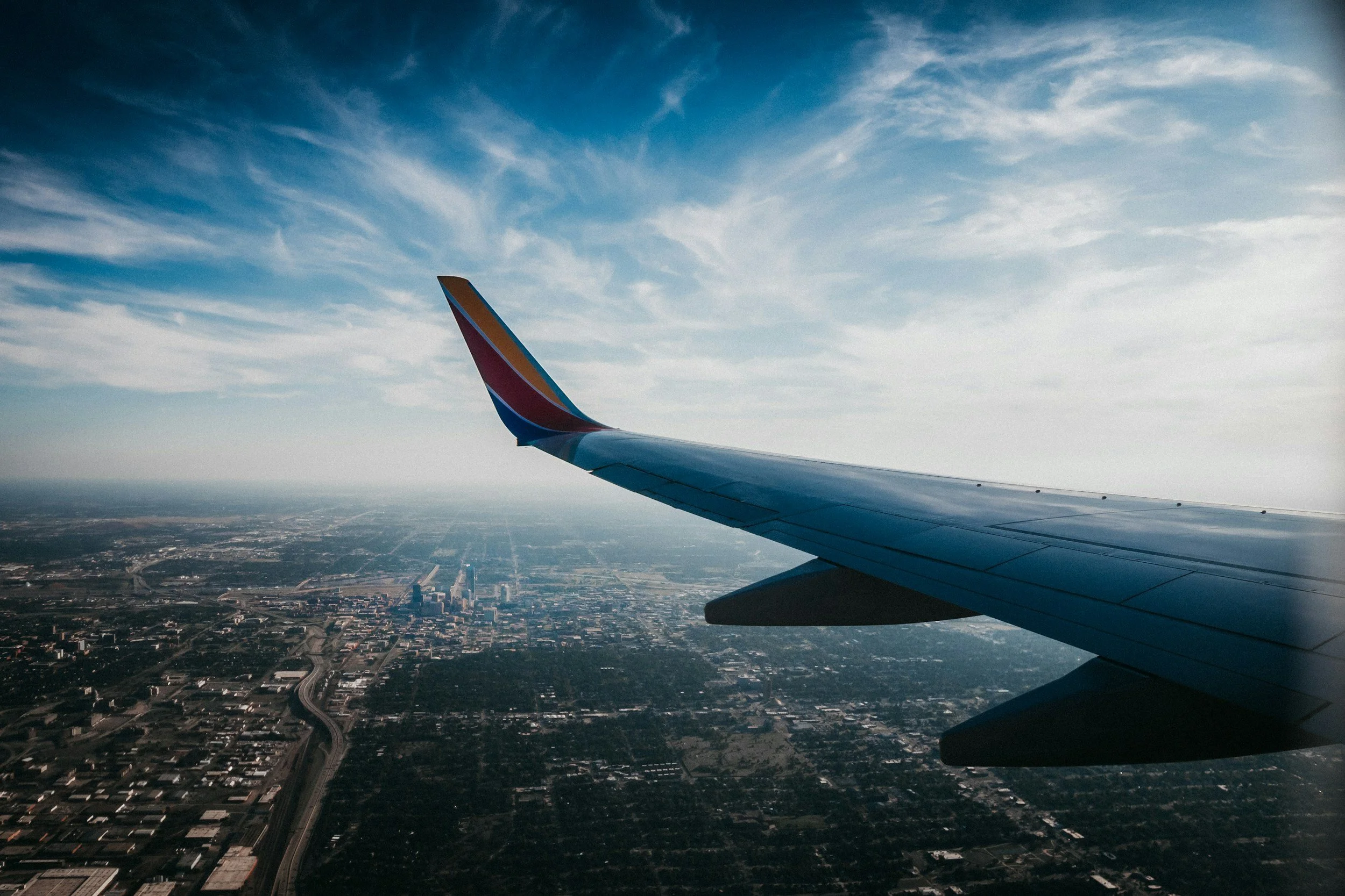 View from an airplane window showing the wing and the city below, with a blue sky and some clouds.