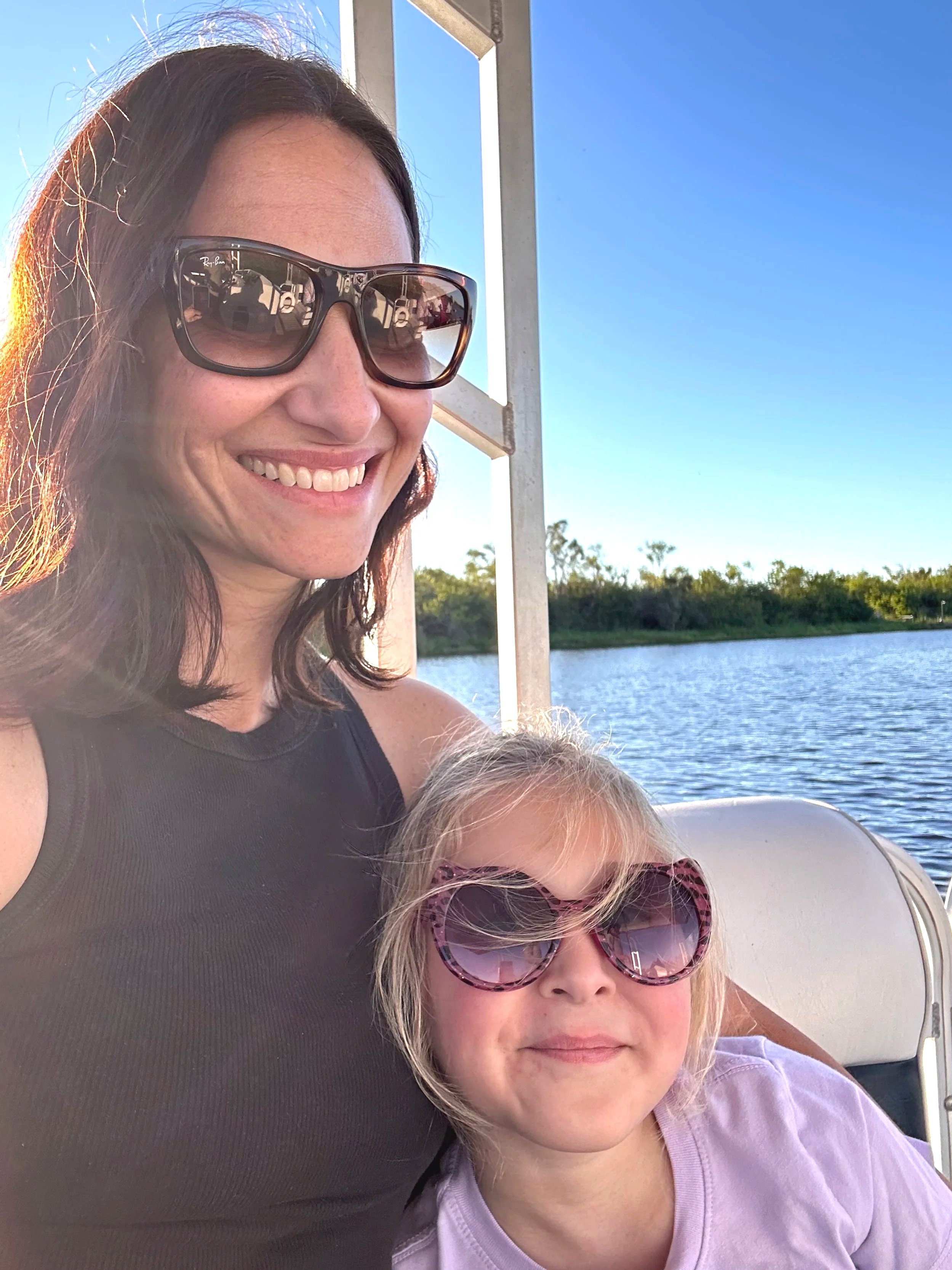 A woman and a girl sitting together outdoors near water, both wearing sunglasses, smiling at the camera with trees in the background on a sunny day.