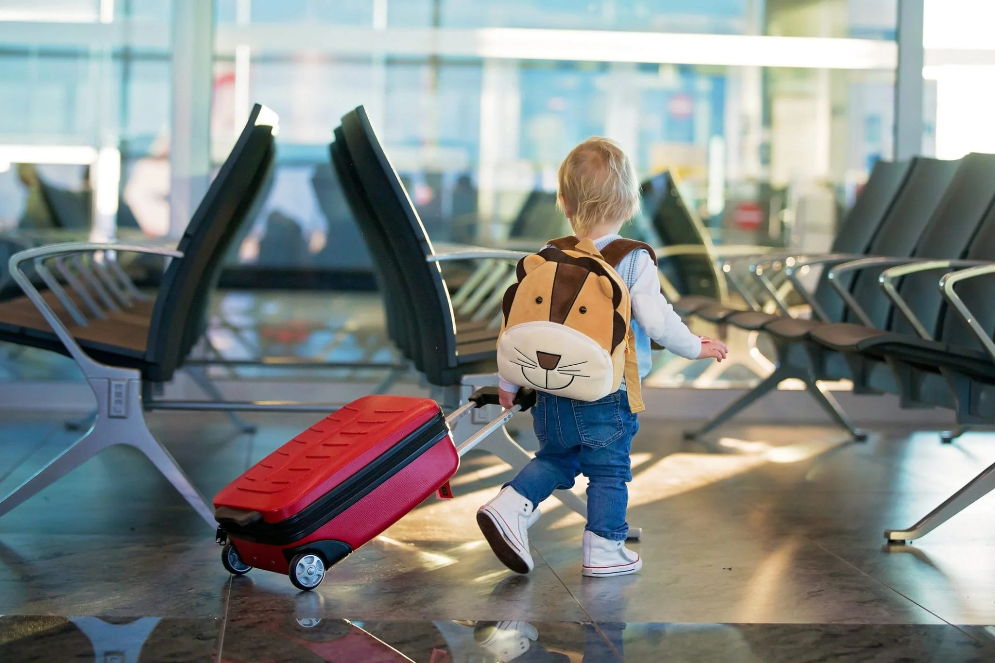 A young child with a lion-themed backpack and white sneakers pulls a red rolling suitcase at an airport waiting area.