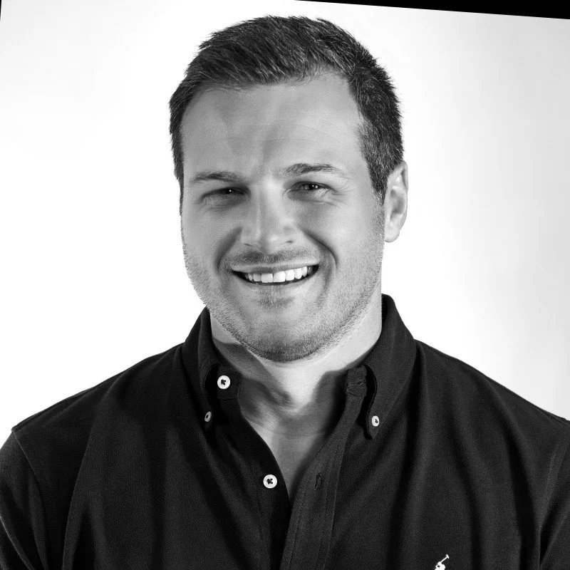 A smiling man with short hair wearing a black collared shirt, looking directly at the camera.