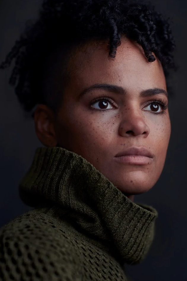 Close-up portrait of a young woman with short curly hair, freckles, and a contemplative expression, wearing a green knitted turtleneck sweater.