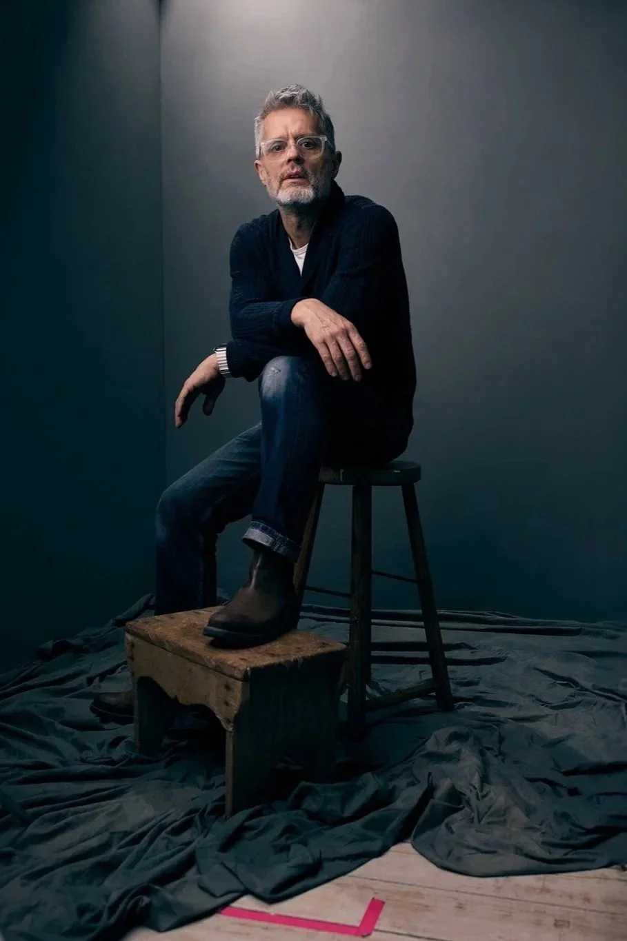 A man with glasses, gray hair, and a beard sitting on a stool, with one foot on a small wooden box, in a studio with a dark background and fabric on the floor.