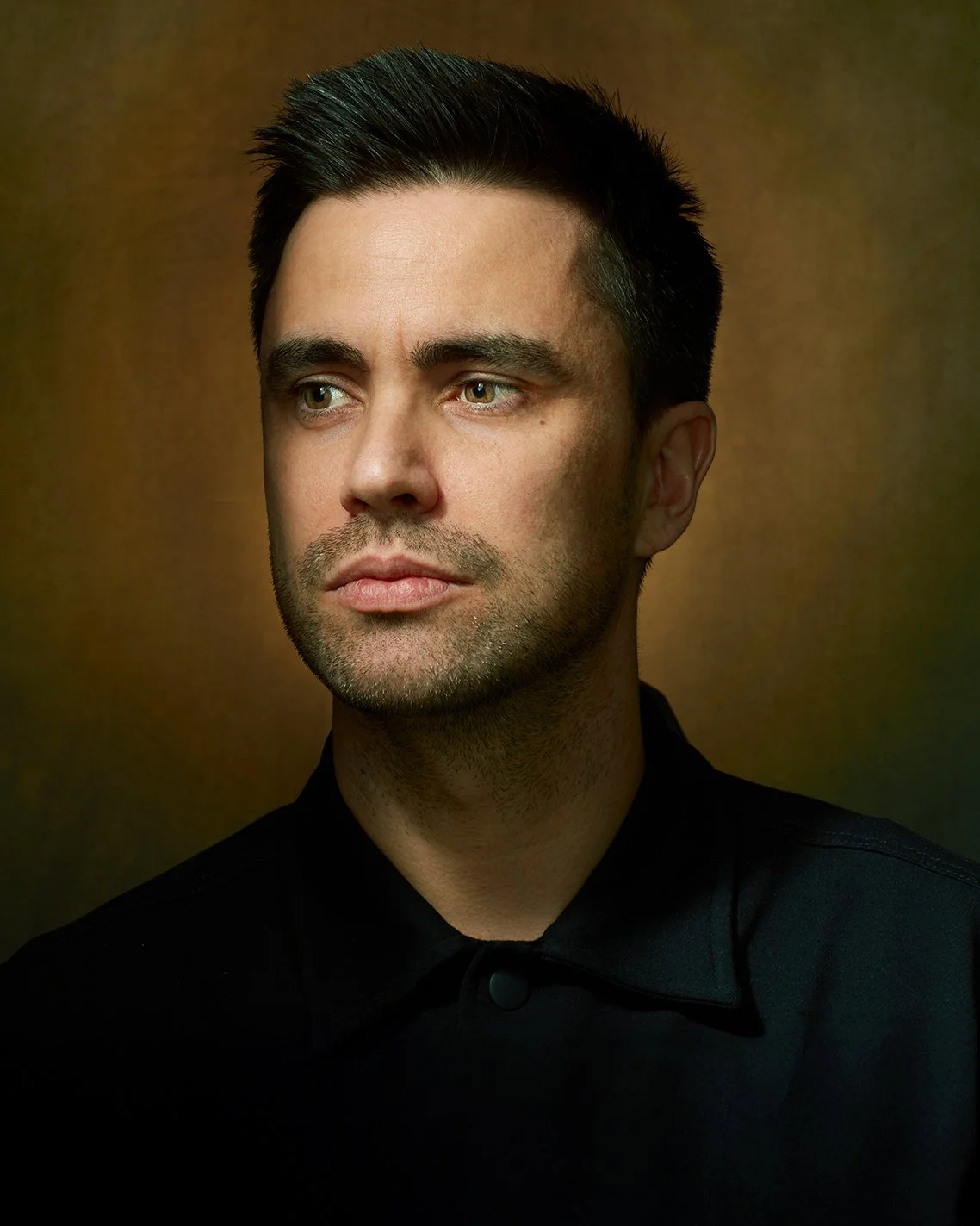 A man with dark hair and light skin wearing a black collared shirt, looking to the side against a dark, softly illuminated background.