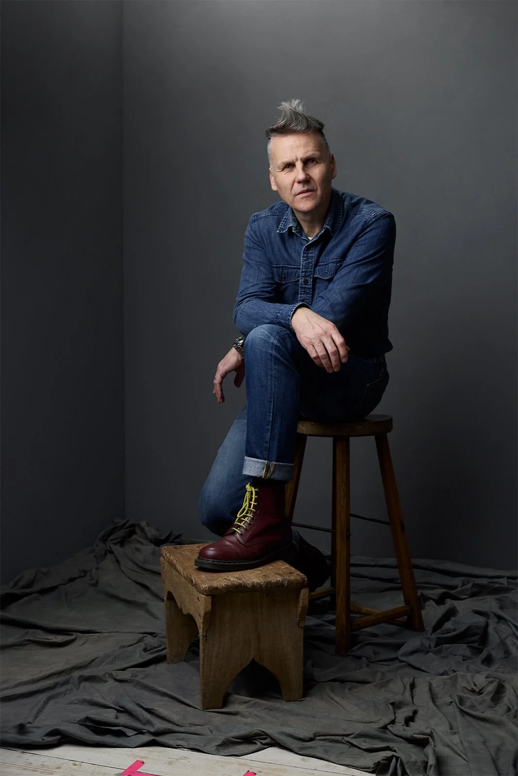 A man with styled gray hair in a blue denim shirt and jeans, sitting on a wooden stool with one leg up, wearing red boots with yellow laces, against a gray wall with a black cloth on the floor.