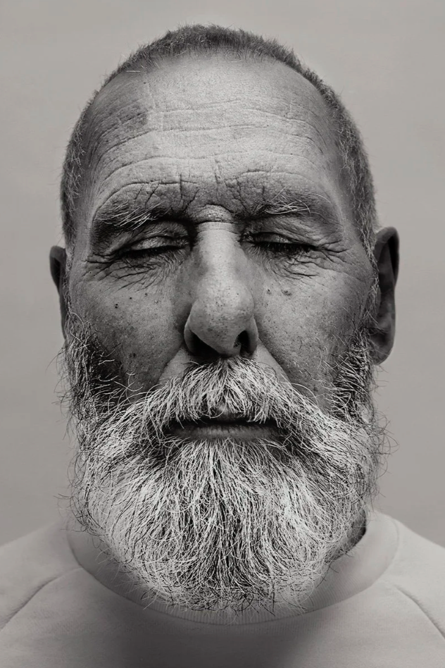 Close-up of an elderly man with closed eyes, gray beard, and wrinkled face, photographed in black and white.