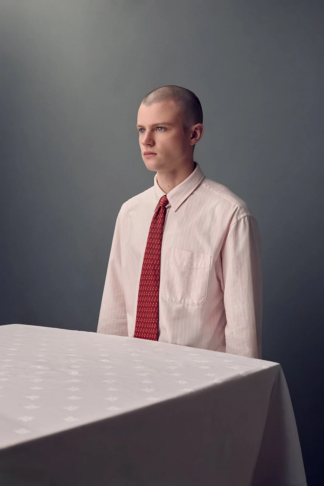 A young man with a shaved head wearing a light pink dress shirt and red tie, standing behind a white table with a patterned cloth, against a plain dark gray background.
