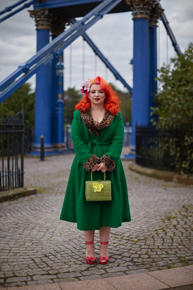 A woman dressed in vintage style with bright orange hair, a green coat with leopard print accents, red polka dot heels, holding a green handbag, standing on a cobblestone path at an amusement park with a roller coaster in the background.