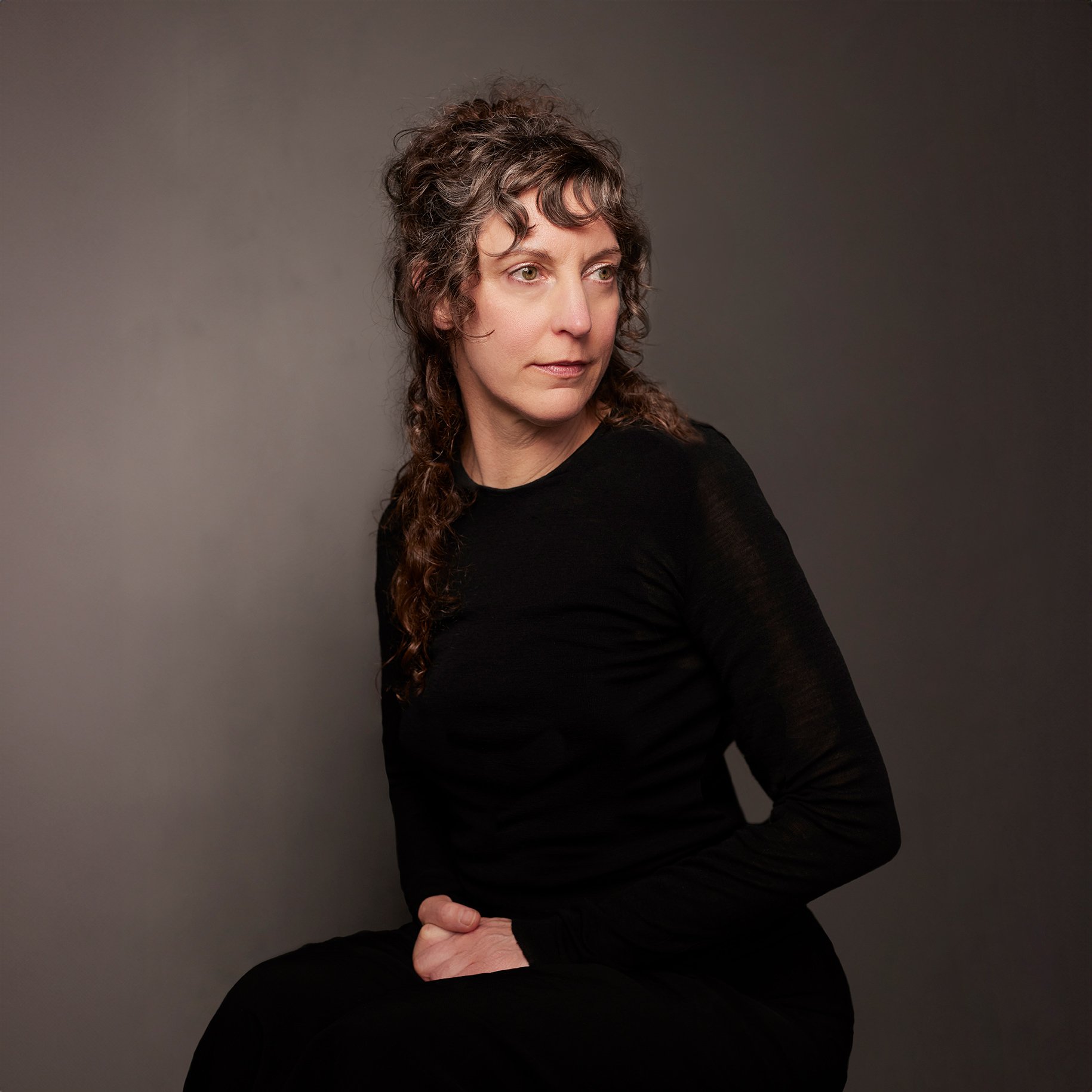 A woman with curly brown hair wearing a black long sleeve top, seated against a dark gray background, looking slightly to her left.