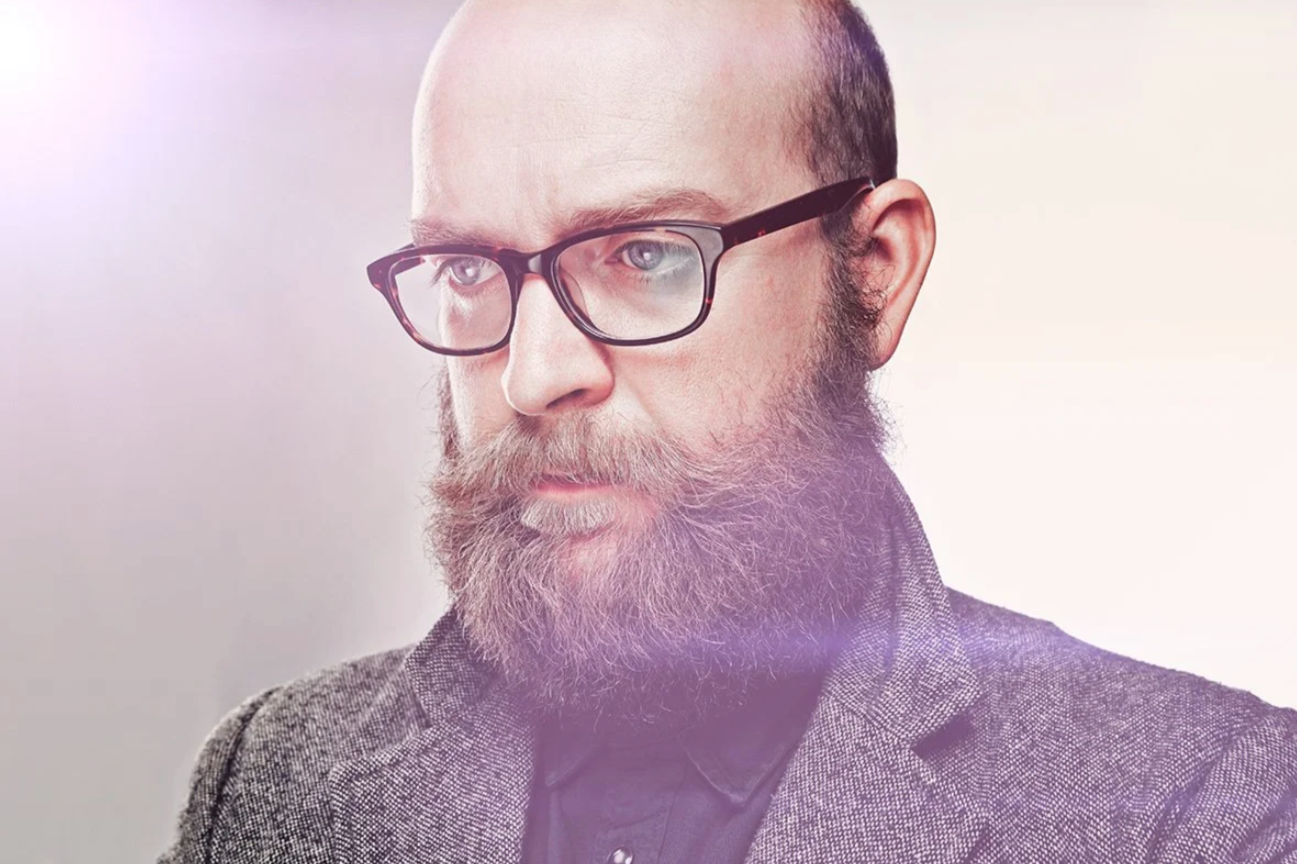 Close-up portrait of a man with glasses, a full beard, and a receding hairline, wearing a gray textured blazer and dark shirt, looking contemplatively to the side.