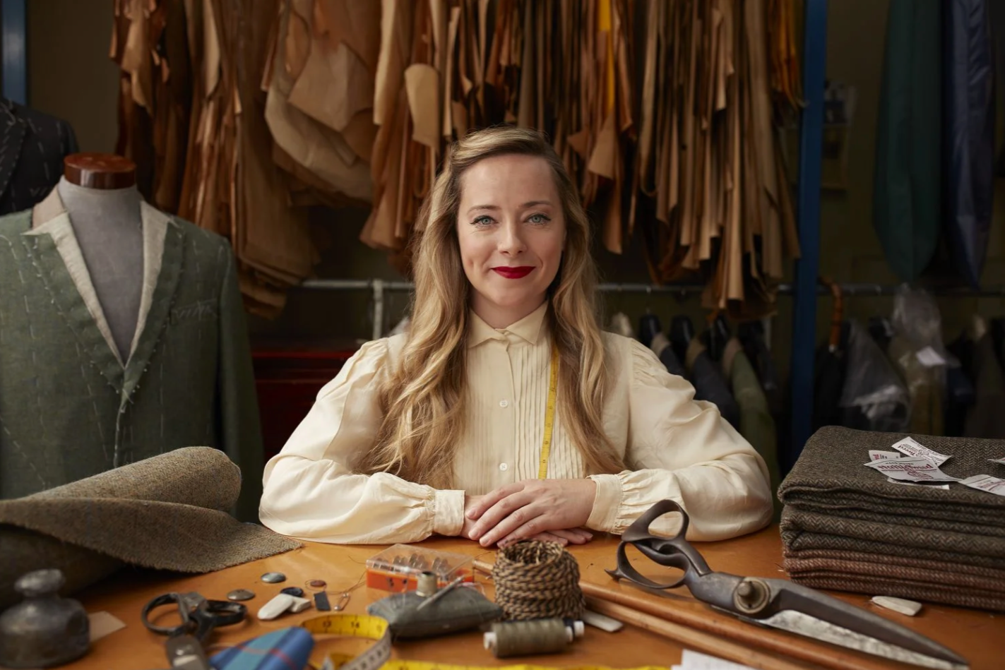 A woman with long blonde hair, red lipstick, and a cream-colored blouse sitting at a work table in a tailor shop surrounded by fabric, scissors, sewing tools, and swatches.