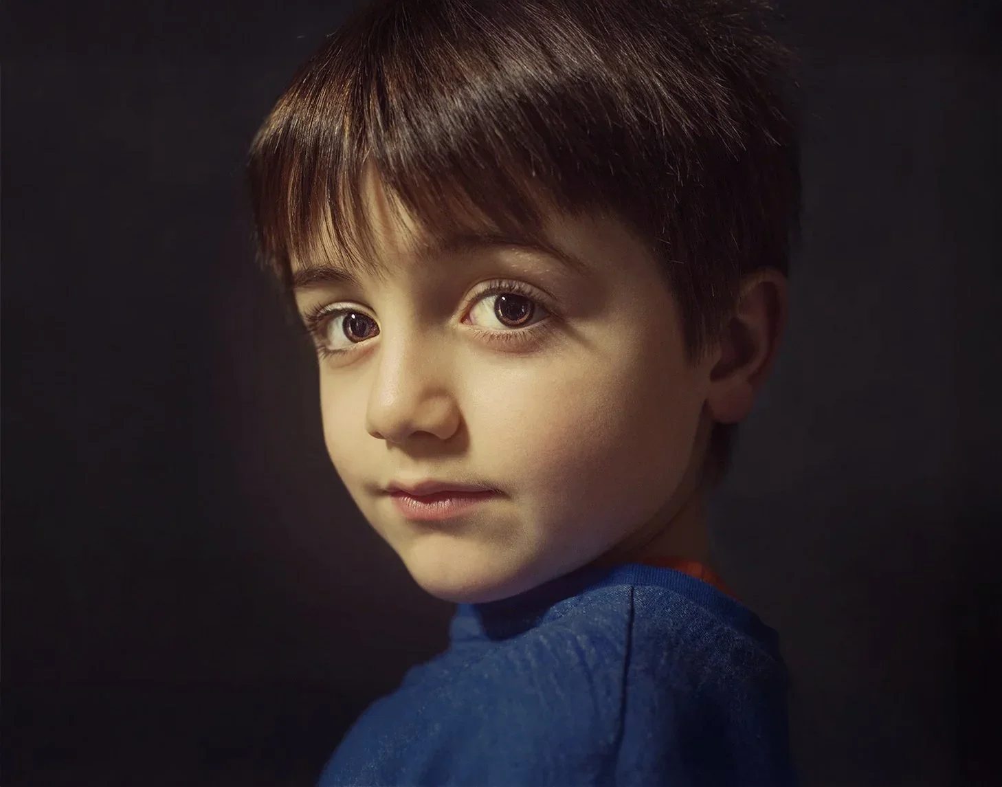 Close-up portrait of a young boy with fair skin, brown hair, and large brown eyes, wearing a blue shirt, against a dark background.
