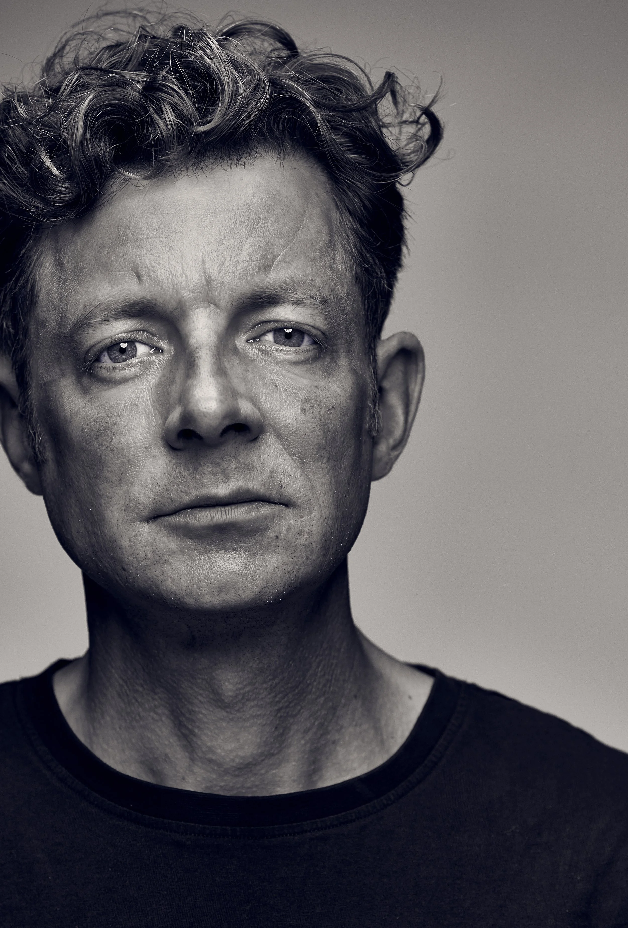 A black-and-white portrait of a middle-aged person with short, curly hair, wearing a dark shirt, looking directly at the camera with a serious expression.