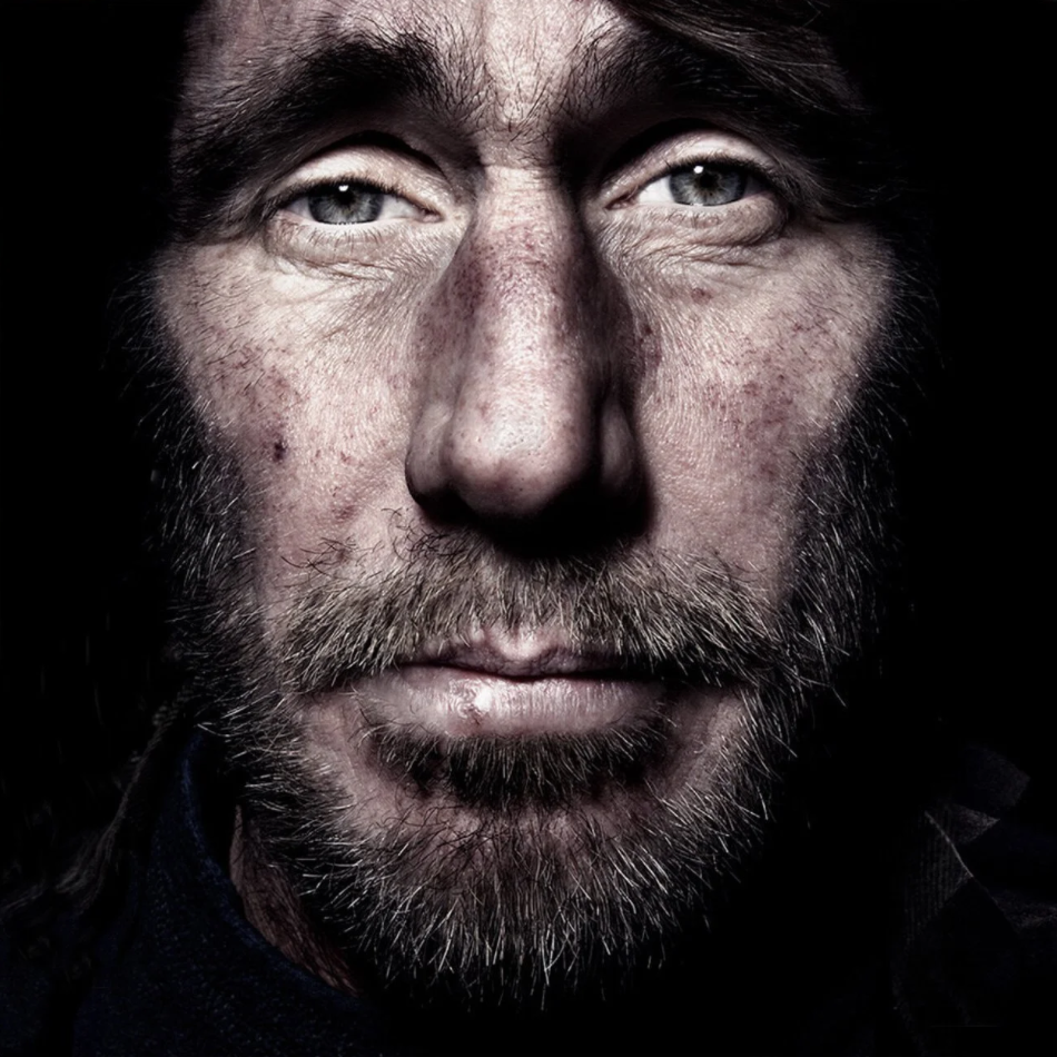 Close-up of a man's face with a beard, showing detailed skin texture, light eyes, and dark hair against a black background.