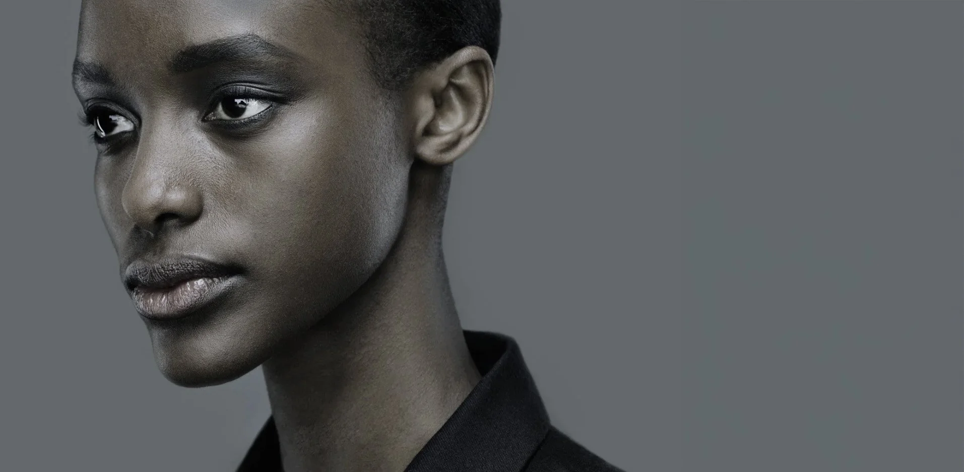 Close-up of a young woman with dark skin, short black hair, wearing a black blazer, against a plain gray background.
