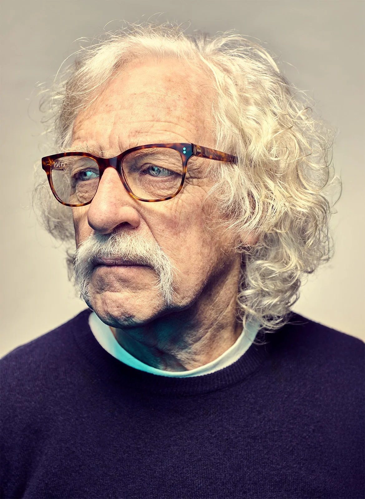Close-up of an elderly man with curly gray hair and a mustache, wearing tortoiseshell glasses and a navy sweater, looking to the side against a neutral background.