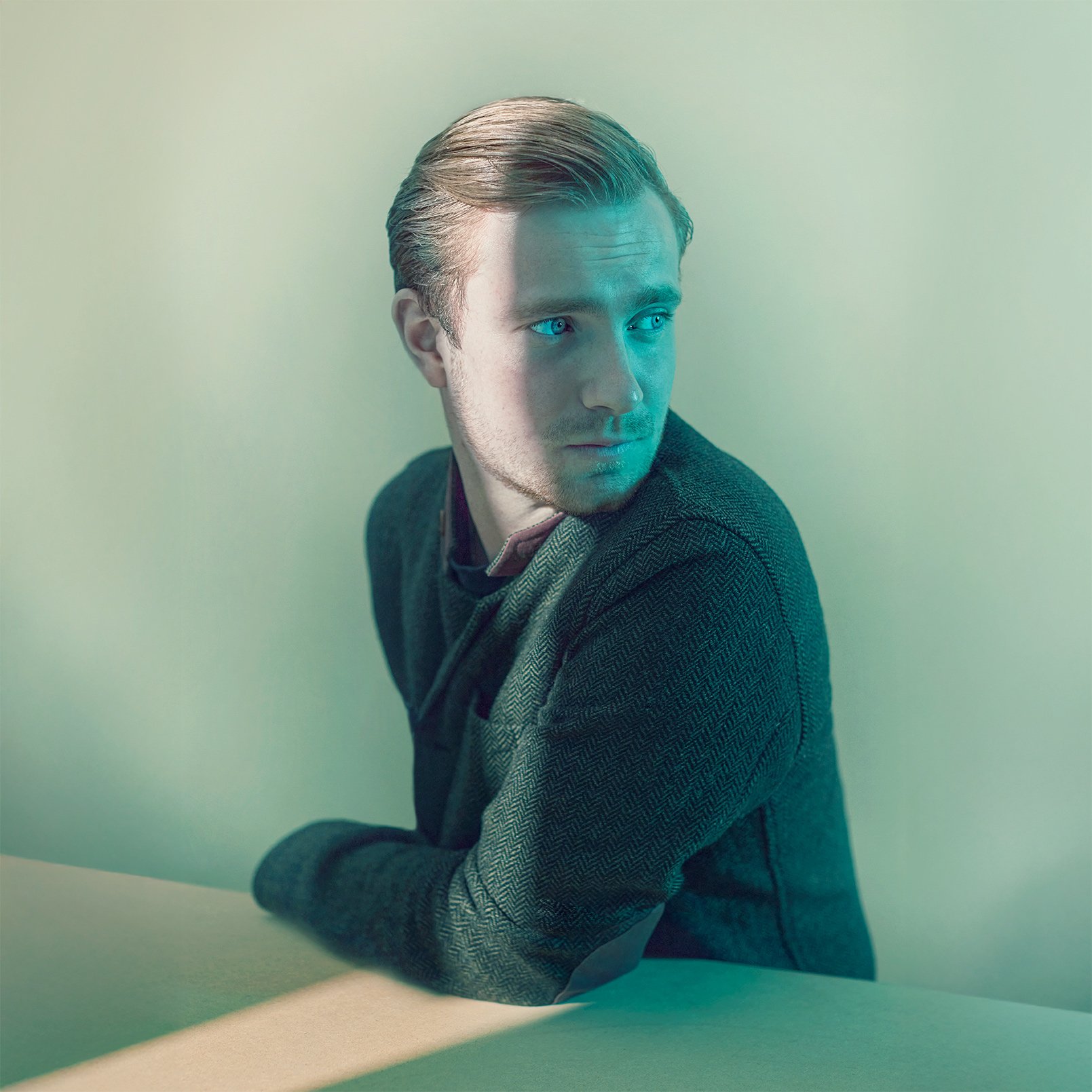 A young man with brown hair and blue eyes looking to the side, sitting at a table with a neutral background.