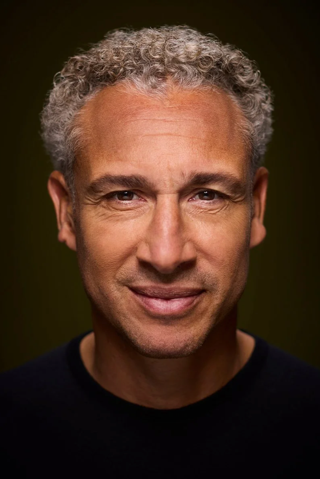 Close-up portrait of a middle-aged man with curly gray hair, smiling slightly, wearing a black shirt against a dark background.