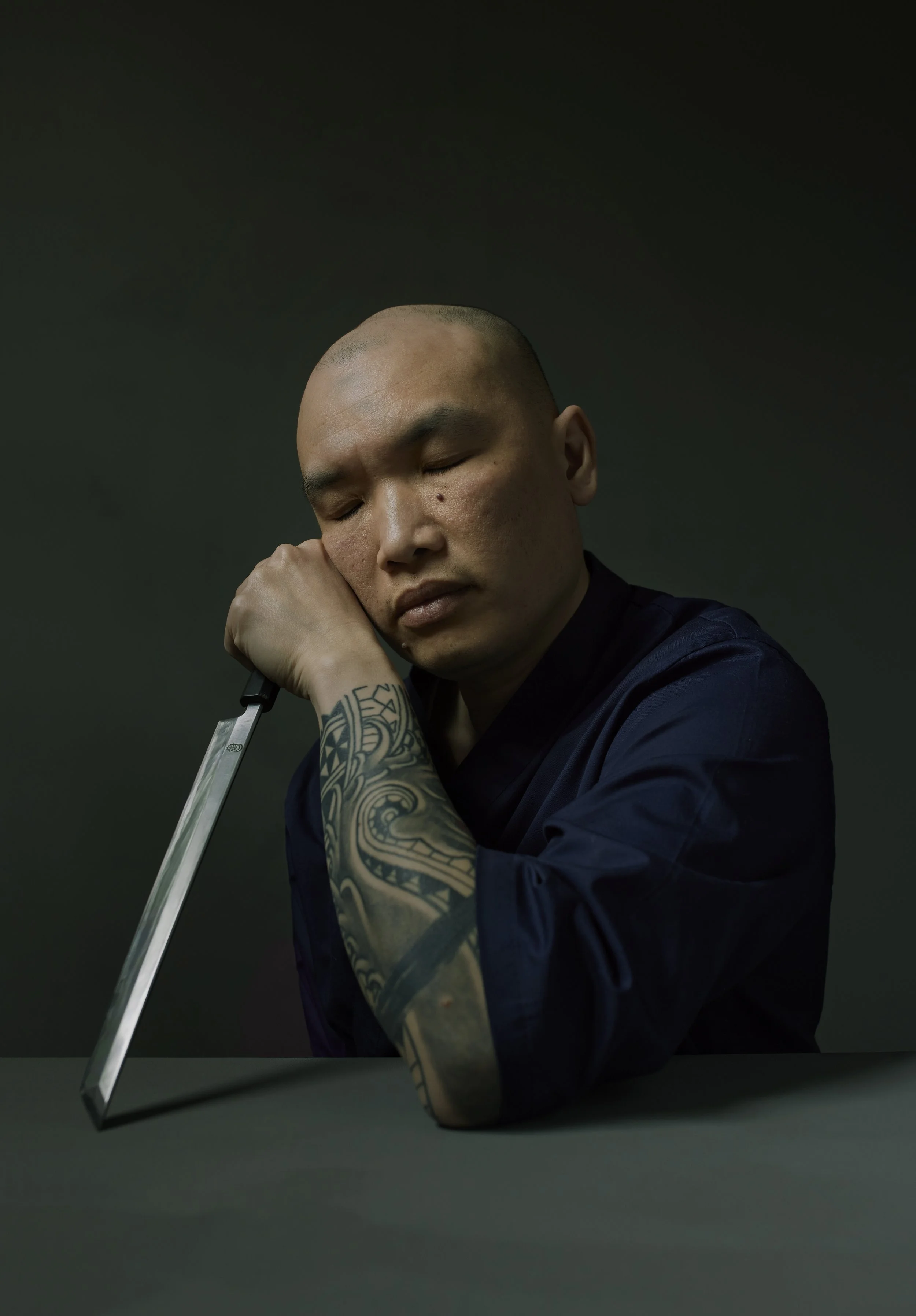 Award winning portrait of Leung Sushi Chef from Sushi Riot , shaved head and tattoos, resting his head on his hand and holding a large knife with his eyes closed against a dark background.