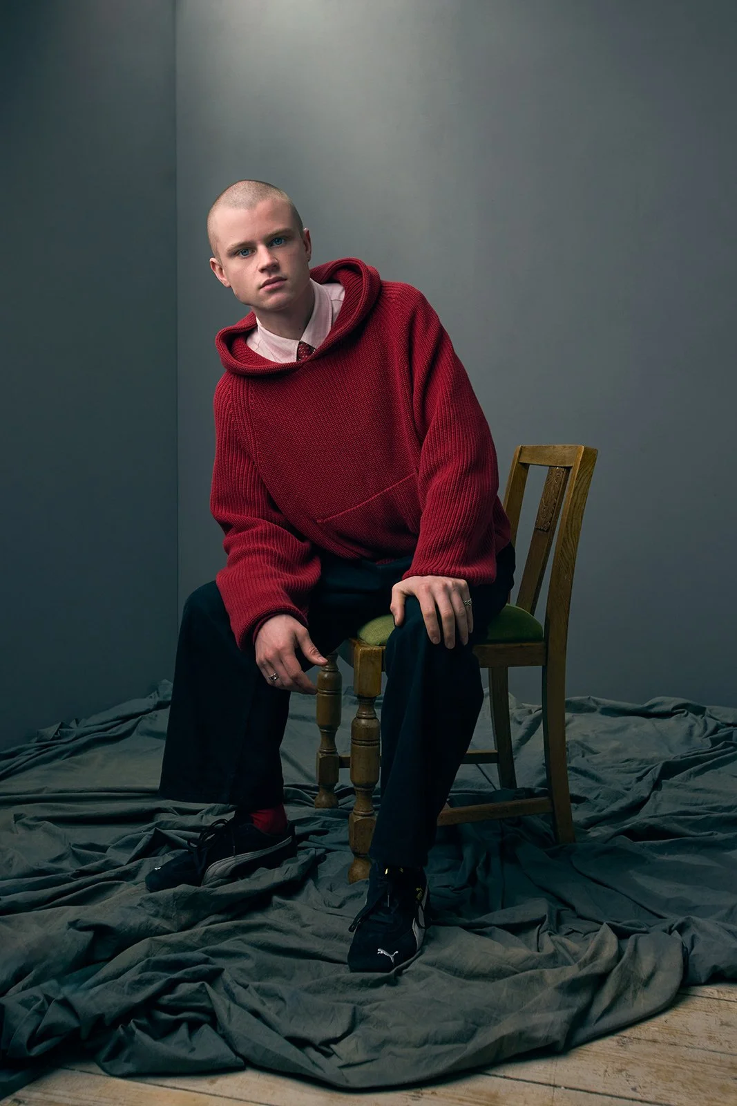 A young man with a shaved head wearing a red hoodie, beige shirt, black pants, and black sneakers, sitting on a wooden chair in a plain room with gray walls and a crumpled gray sheet on the floor.