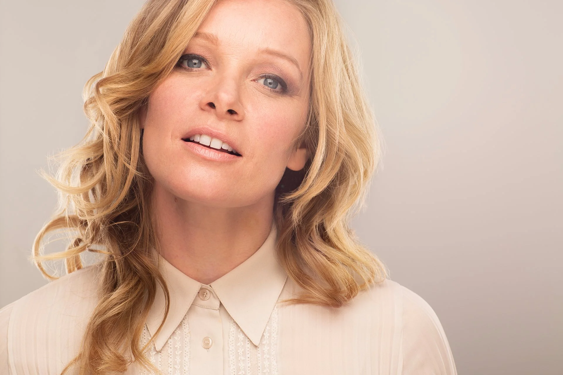 A woman with blonde hair styled in loose waves, wearing a cream-colored blouse with a pointed collar. She has light makeup with neutral tones, light eyeshadow, and a soft smile, standing against a plain, light background.
