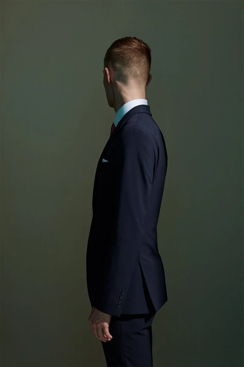 Side view of a man in a dark suit facing away from the camera, against a plain green background.