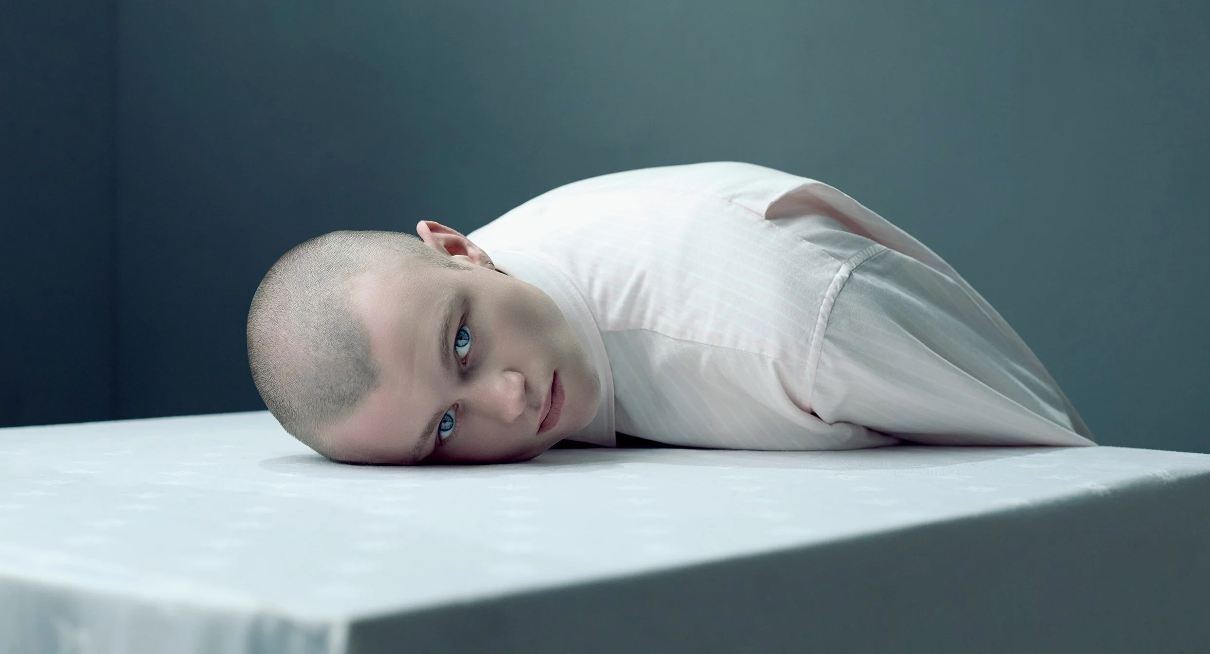 Person with a shaved head lying on a bed, looking up, wearing a white shirt, with a serious expression, in a minimalist room.