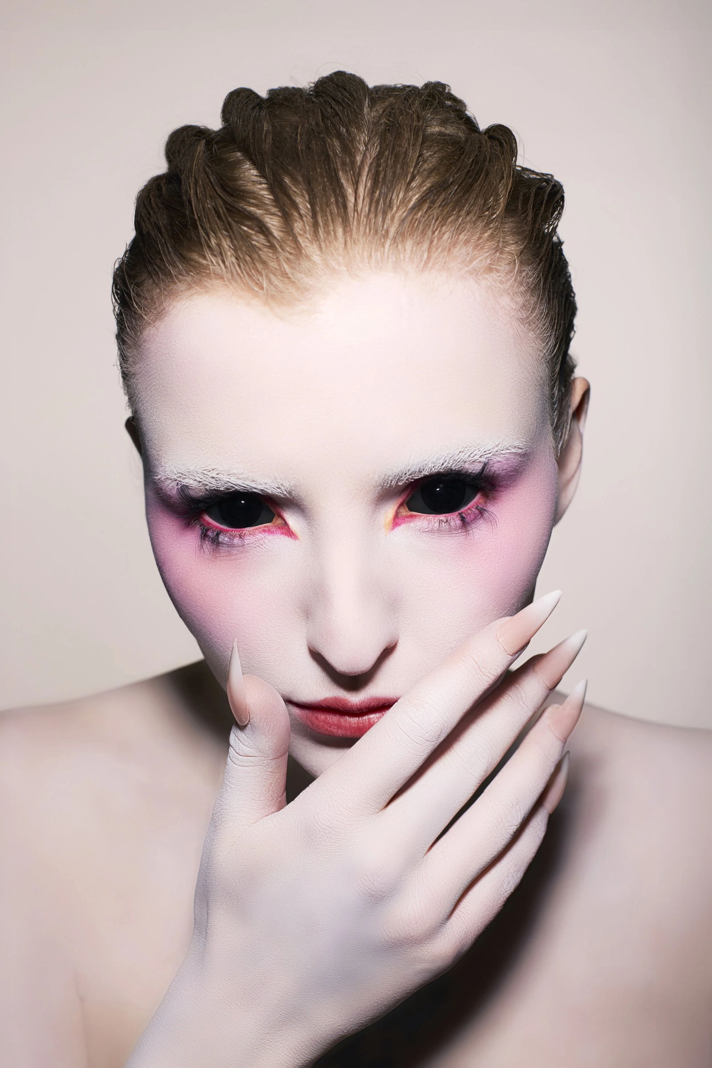 A woman with avant-garde makeup, pale skin, white eyeshadow, pink blush, and long manicured nails poses with her hand near her face against a neutral background.