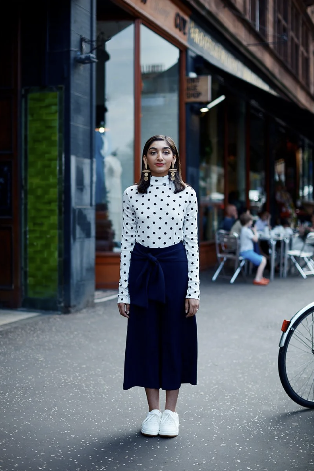 A young woman with dark hair wearing a white polka dot long sleeve shirt, navy blue high-waisted skirt with a tied bow, and white sneakers, standing on a city sidewalk in front of a glass storefront with other people sitting outside a cafe in the bac