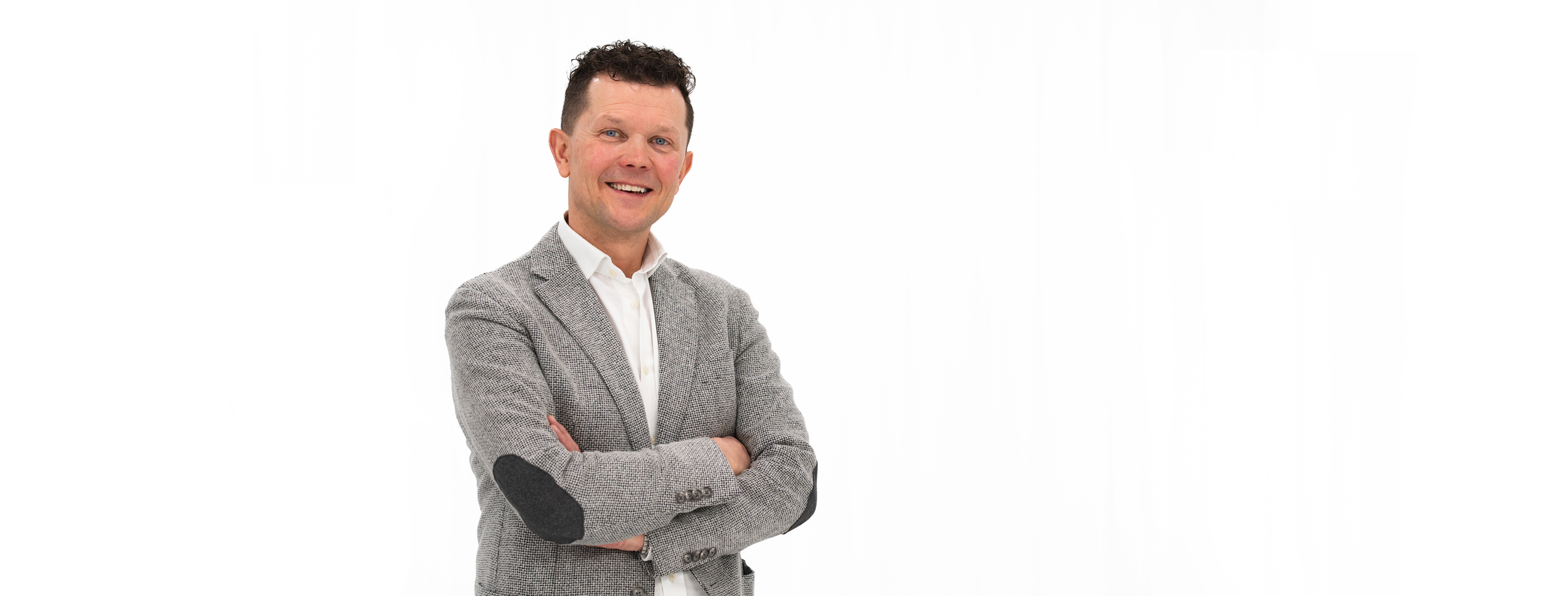 Dr. Roman Straßl trägt einen grauen Blazer mit Ellbogenpatches und ein weißes Hemd, steht vor weißem Hintergrund und lächelt.