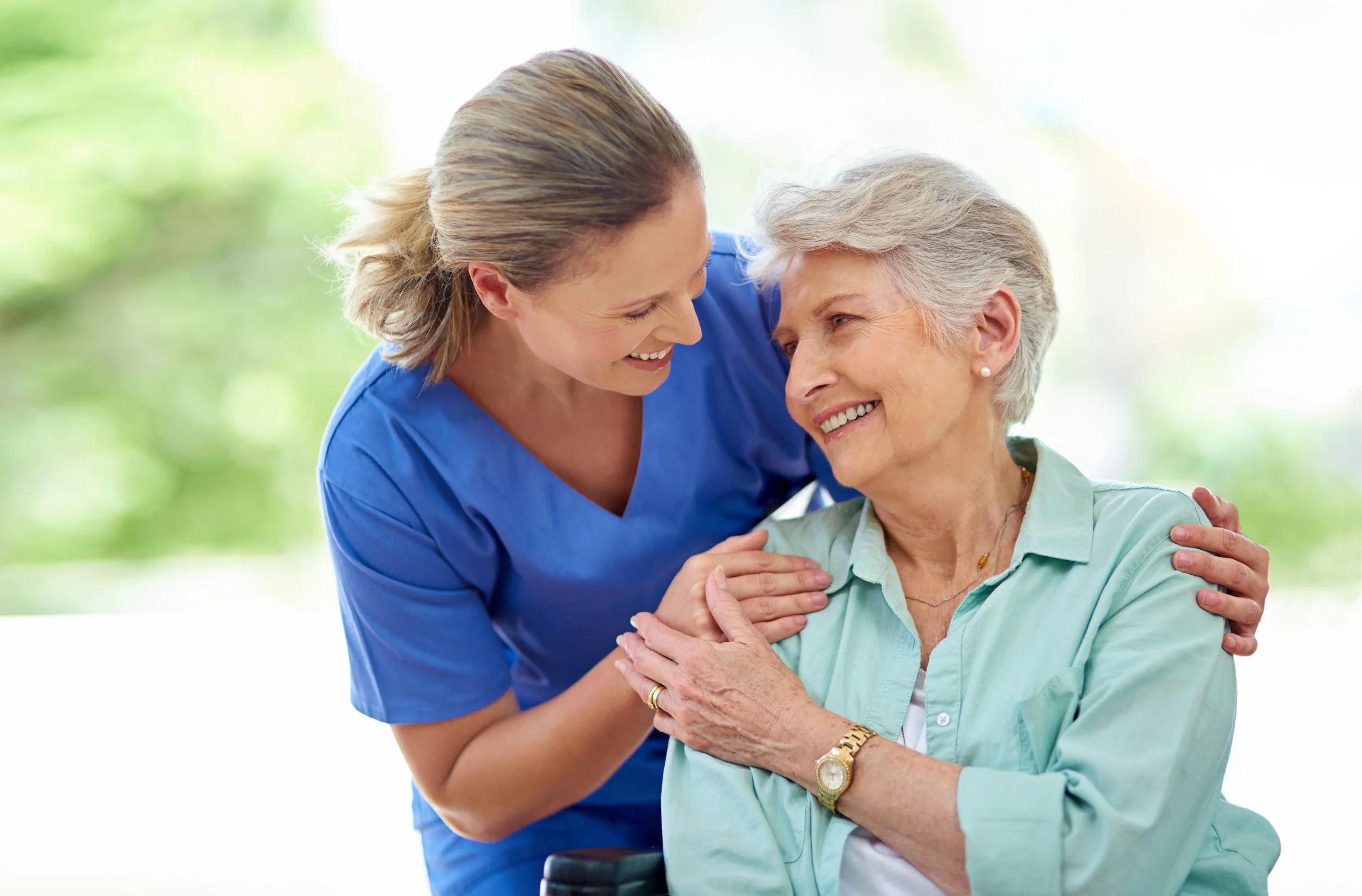 A healthcare worker, dressed in blue scrubs, is smiling and holding hands with an elderly woman who is sitting in a wheelchair. The elderly woman is wearing a light green blouse and has short gray hair, and she is smiling back at the healthcare worker.